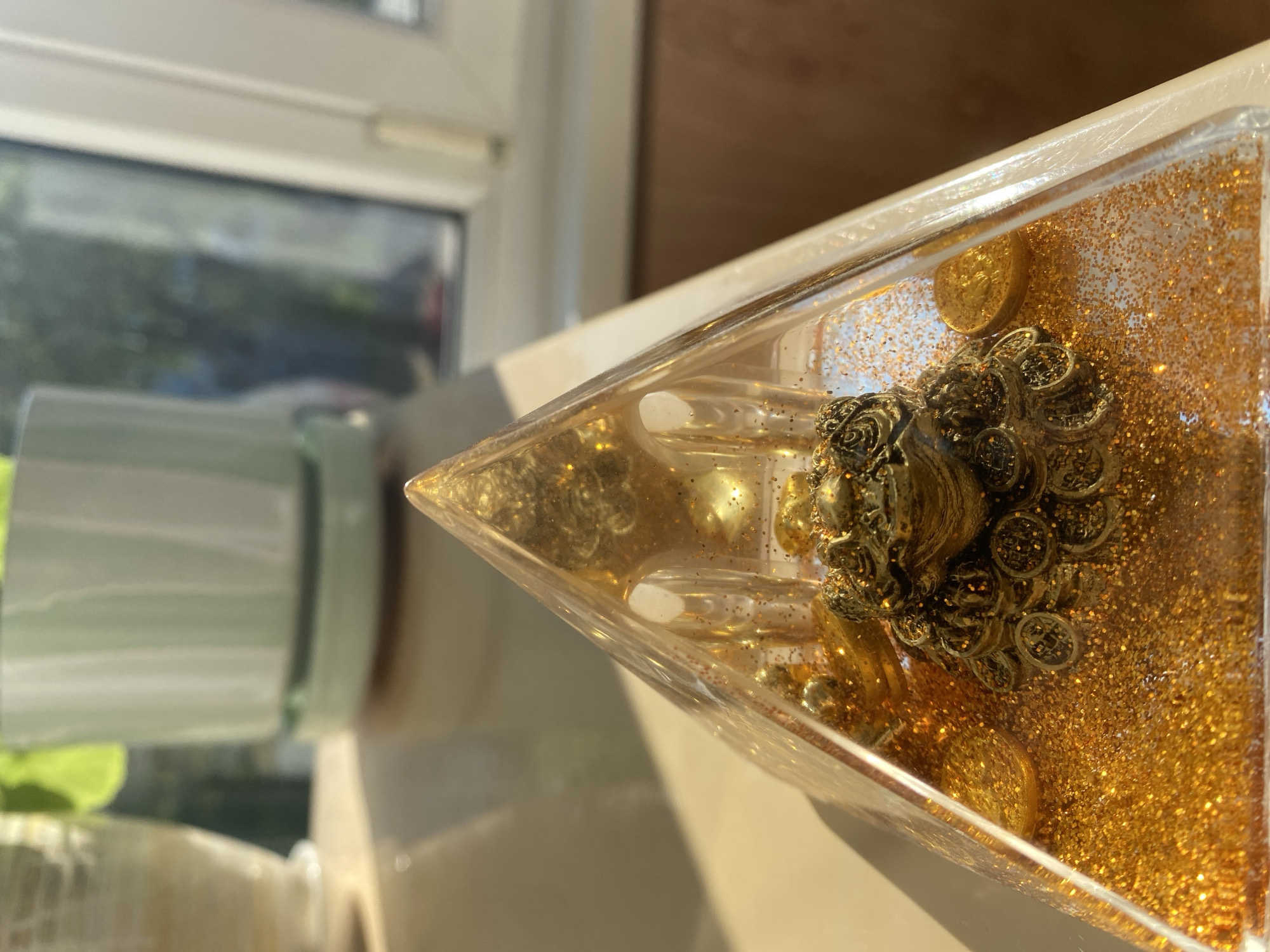 A close-up, eye-level shot shows a golden, three-legged toad figurine positioned inside a clear, pyramid-shaped object filled with gold glitter and coin-like embellishments. The figurine is detailed, with intricate textures on its body and what appear to be coins stacked around its base. The pyramid is illuminated by sunlight, casting shadows and highlighting the glitter. In the blurred background, a light-colored plant pot with a green plant is visible, along with a window that shows a reflection of trees and a dark silhouette of a person. The surface beneath the pyramid is smooth and reflective, with a light tan hue. There are no people or animals present in the immediate scene. The location is not discernible from the image.