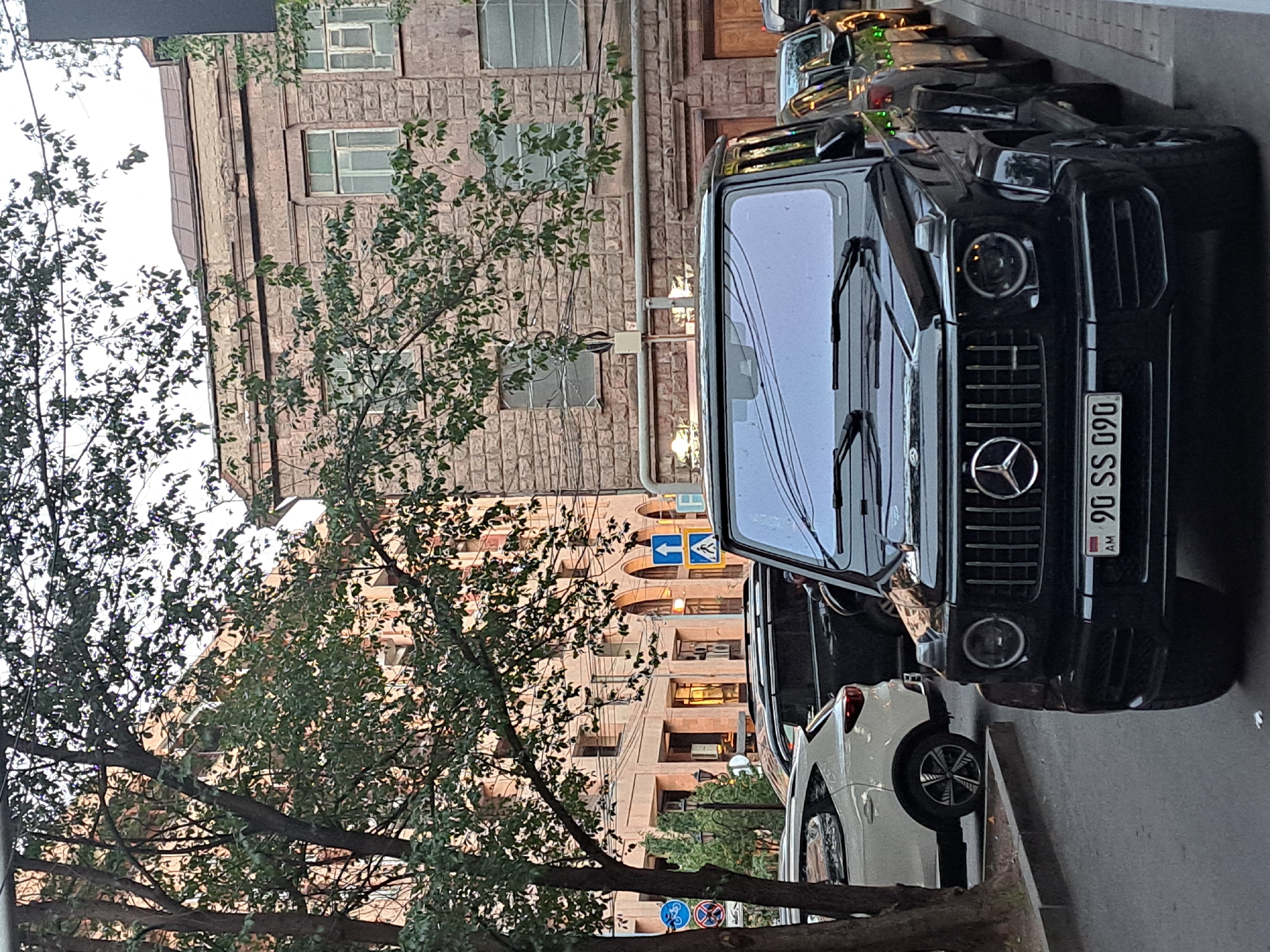The image shows a black Mercedes-Benz G-Class parked on a city street with buildings in the background. The vehicle has a license plate that reads "90 SS 090" and the "AM" indicates the license plate is from Armenia. The buildings appear to be made of stone, and there are trees with green leaves that are in the foreground. There are a few traffic signs visible on posts in the background. The sky appears overcast. Based on the style of architecture and license plate on the vehicle, it is likely this photo was taken in Yerevan, Armenia.