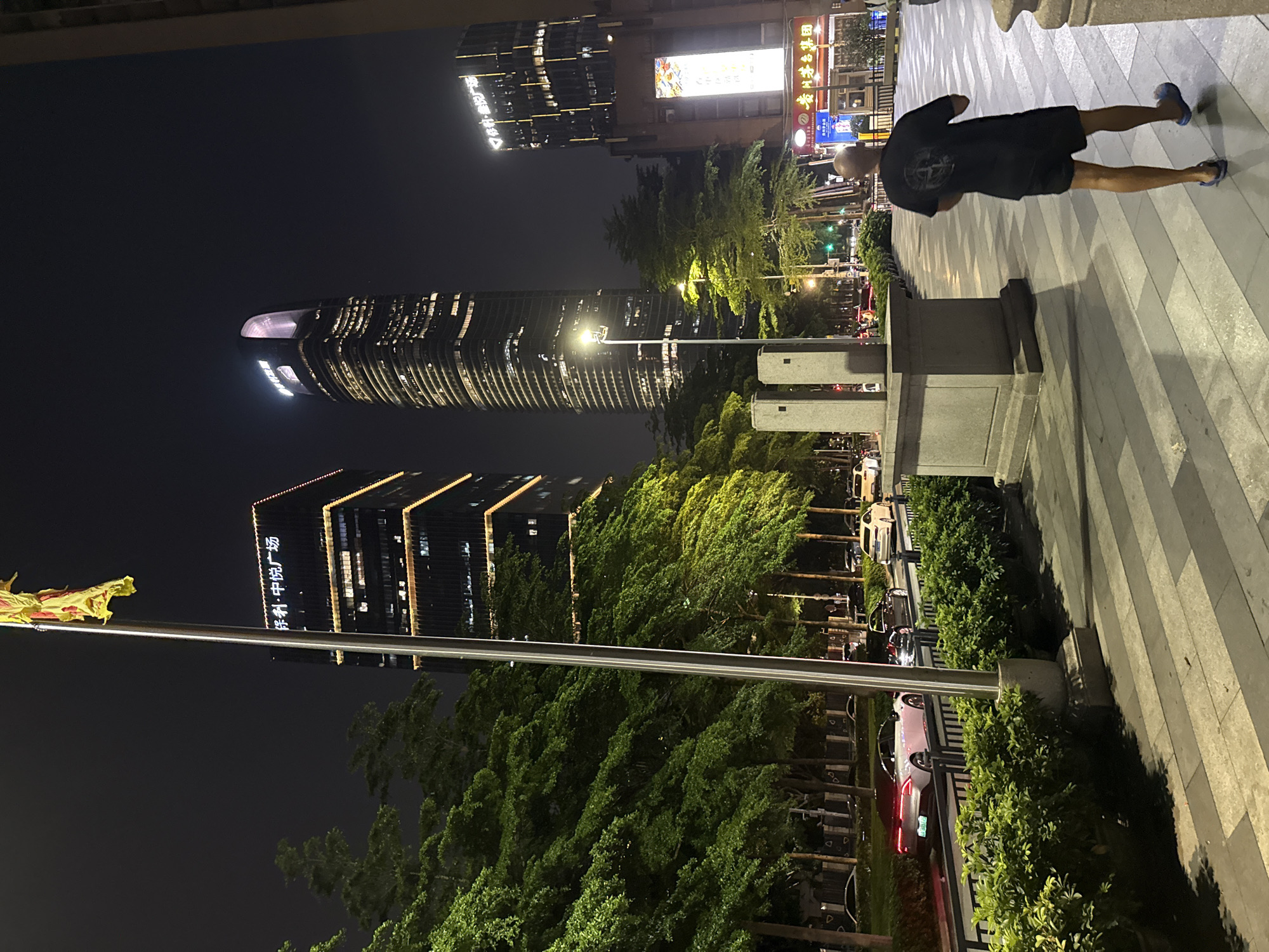 The image captures a vibrant urban street scene at night in Haizhu District, China. The primary subject in the foreground is a person, likely male, seen from behind. He is wearing a dark t-shirt and dark shorts, with light blue flip-flops on his feet, standing on a grey and white paved sidewalk.
The background is dominated by several brightly lit modern skyscrapers against a dark night sky. The most prominent is a tall, curved building with numerous illuminated windows, showcasing its unique architecture. To its left, another large building features distinctive horizontal and vertical illuminated lines, and displays Chinese characters spelling "保利·中悦广场" (Bǎolì·Zhōngyuè Guǎngchǎng), which translates to "Poly·Zhongyue Plaza". Further buildings in the distance also contribute to the city's illuminated skyline, with various signs displaying Chinese script, indicating shops or businesses.
The street level features lush green trees and bushes, creating a natural element within the urban landscape. A busy road runs parallel to the sidewalk, with several cars visible, some parked and others driving, their headlights and taillights glowing. Streetlights cast pools of light on the pavement and vegetation. A metal pole in the mid-ground holds a yellow and red flag or banner, adding a touch of color to the night scene. The overall impression is a modern, bustling city center alive with lights and activity after dark.