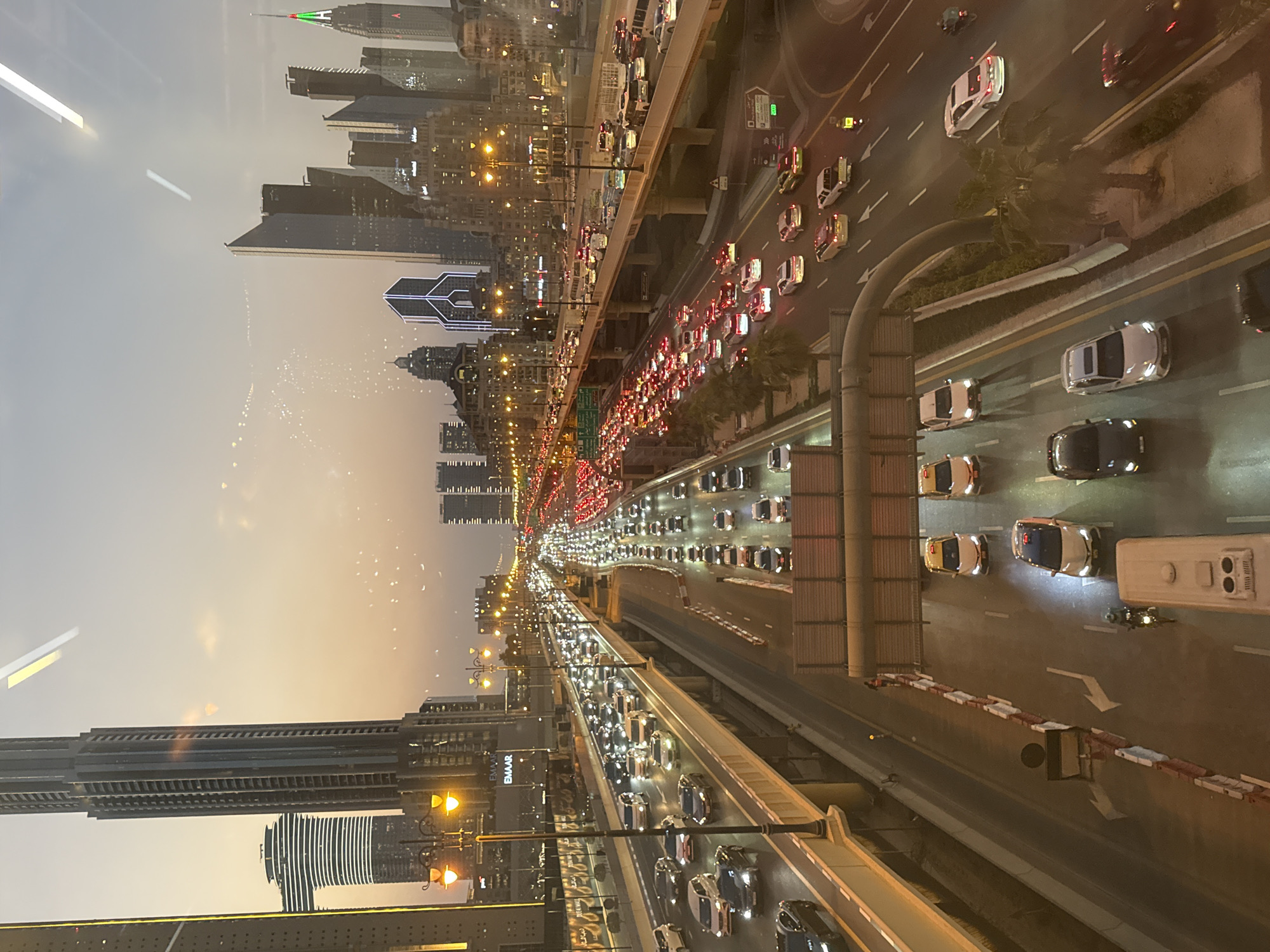 The image depicts a congested highway in Dubai, United Arab Emirates, likely during twilight or early evening. The Sheikh Zayed Road, a major arterial route, is visible, characterized by multiple lanes of traffic filled with cars, taxis, and buses. Towering skyscrapers, characteristic of Dubai's skyline, flank the highway, with illuminated windows and architectural lighting indicating the urban environment. The sky above is a gradient of orange and gray, suggesting either sunrise or sunset. The highway itself is well-lit by streetlights and the headlights and taillights of the vehicles. Barriers and road signs are present, guiding the flow of traffic. The overall impression is one of a bustling, modern city at a busy time of day.