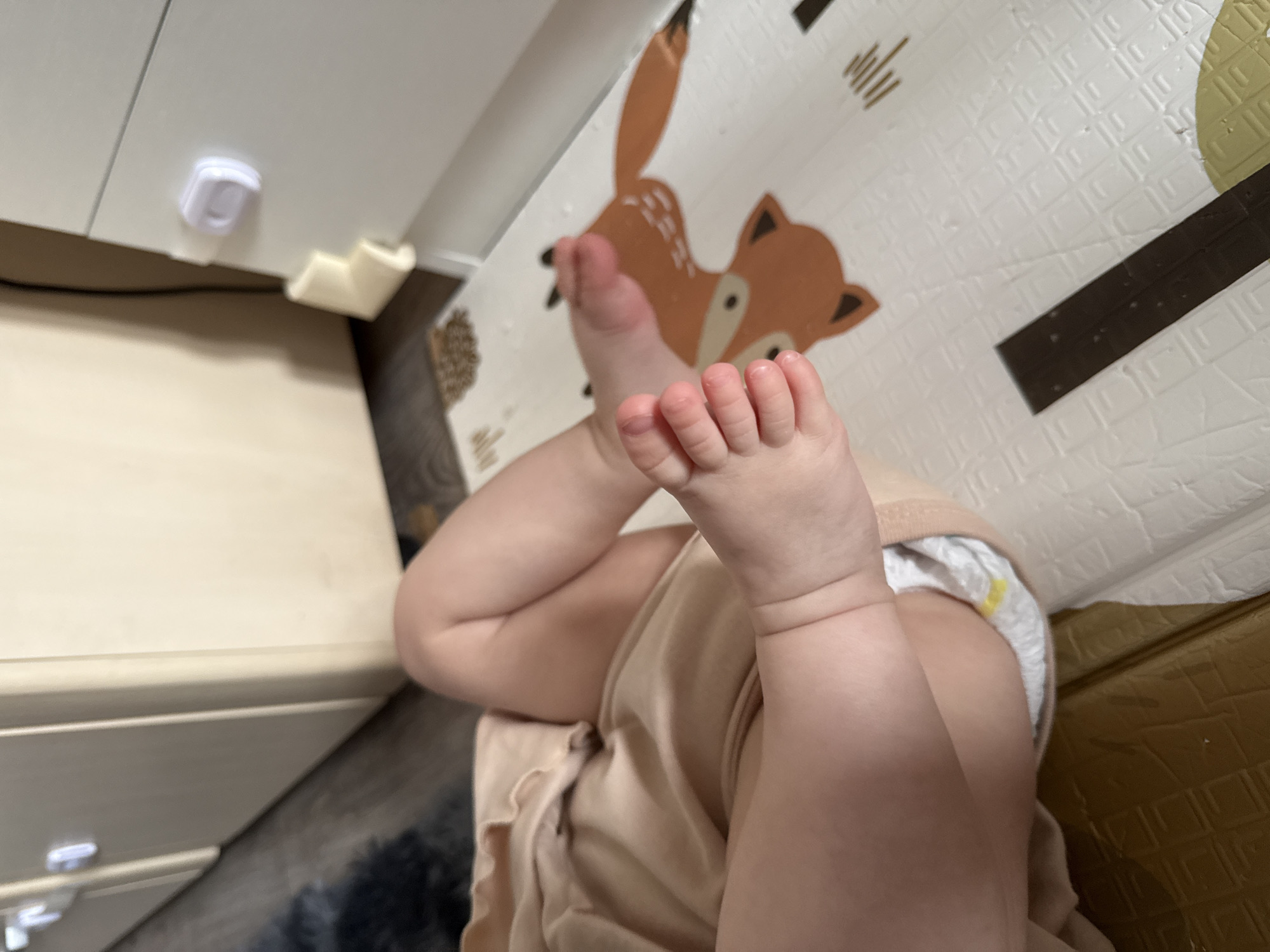 A baby is lying on a playmat. The baby is wearing a diaper and a beige outfit. The baby's legs are raised in the air, with its feet pointed towards the viewer. The playmat has a pattern of animals and geometric shapes. A piece of furniture is visible in the background. There is no evidence to suggest that the image was taken in Yakutsk, Russia.
