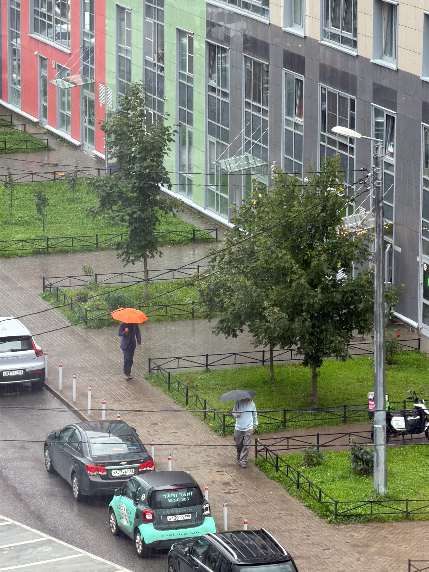 An overhead shot captures a rainy day scene in front of a modern apartment building with a facade of red, light green, and grey panels. A wet brick-paved walkway runs along the building, bordered by a grassy area with small trees and black metal fencing.
On the walkway, two individuals are visible. The first, closer to the building, is walking with an orange umbrella, wearing dark clothing. Further down the path, a second person walks with a grey umbrella, dressed in a light blue hoodie and grey pants.
Several cars are parked along the walkway. A white SUV is parked on the left, its rear facing the viewer. Behind it, a dark grey sedan is parked. To the right of the sedan, a small teal and black smart car, identified as a Yandex.Eda delivery vehicle by its markings, is parked. A white delivery scooter with a large black box on the back is parked further down the walkway near a lamppost.
The overall environment suggests a residential area in Saint Petersburg, Russia, experiencing inclement weather. The wet surfaces and umbrellas indicate active rainfall.