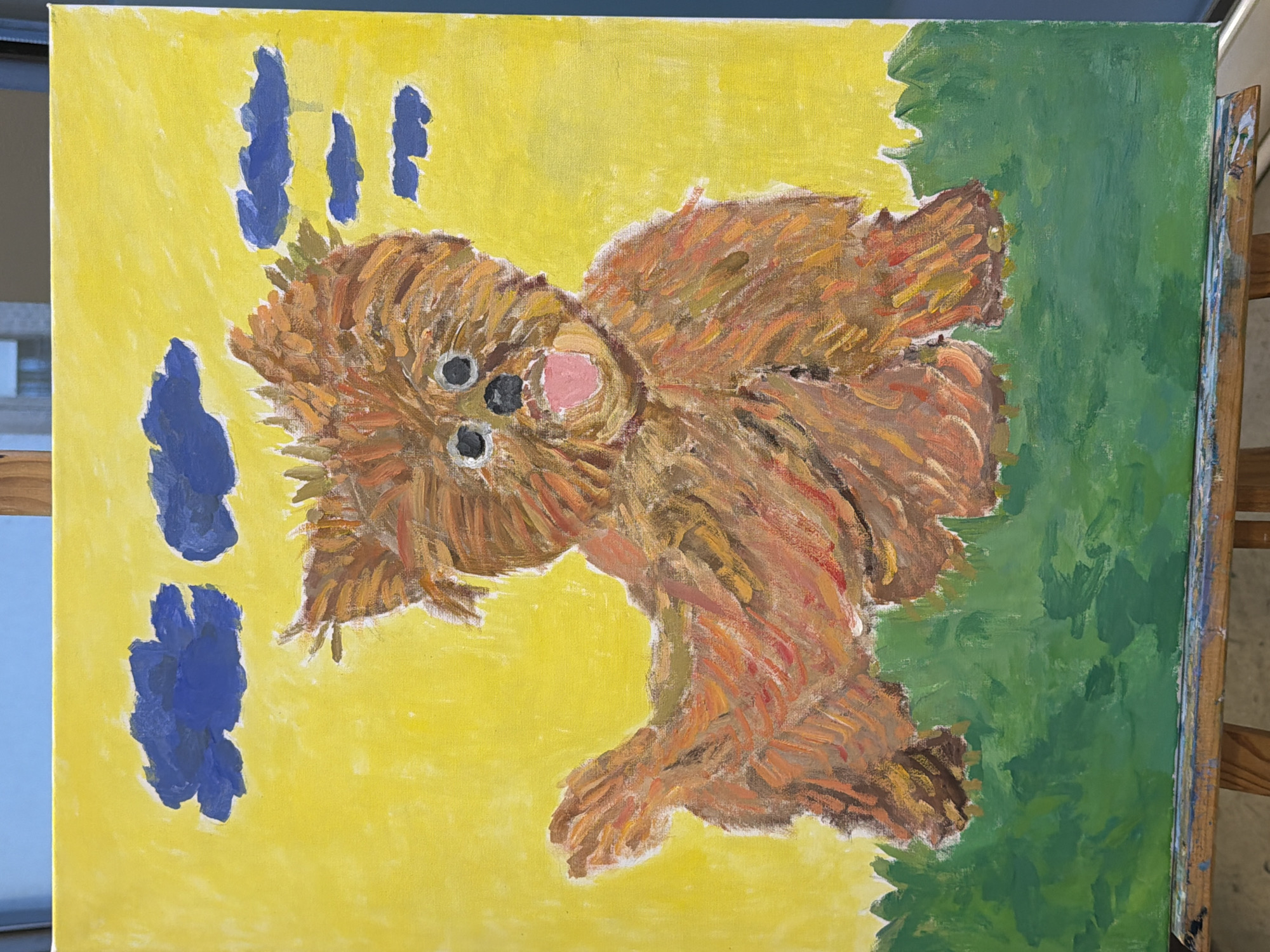 This is a painting of a brown, fluffy dog with its tongue sticking out, sitting in a grassy field under a yellow sky with blue clouds. The dog is the central subject, depicted with thick, textured brushstrokes that give it a shaggy appearance. Its eyes are wide and black, and its ears are perked up. The foreground is a vibrant green, representing grass, while the background is a bright yellow, suggesting a sunny day. The blue clouds are scattered across the top of the canvas. The painting is displayed on a wooden easel. There is no discernible location in the city of Elche/elx, Spain depicted in the image.