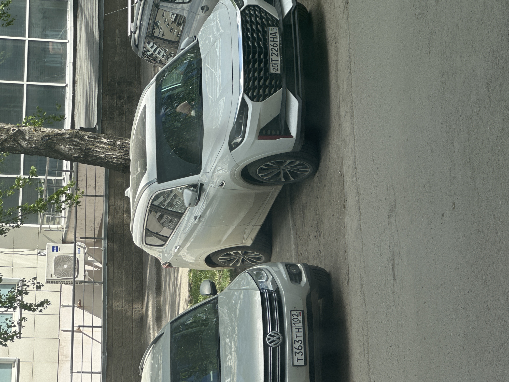 The image shows a street scene in Unknown, Unknown. A white Volkswagen with license plate T363TH102 and a white car with license plate 20 T226HA02 are parked side by side on a paved street. To the left of the cars is a building with a large window and a tree partially obscuring the view. An air conditioning unit is mounted on the side of the building. The environment is sunny, suggesting it is daytime.