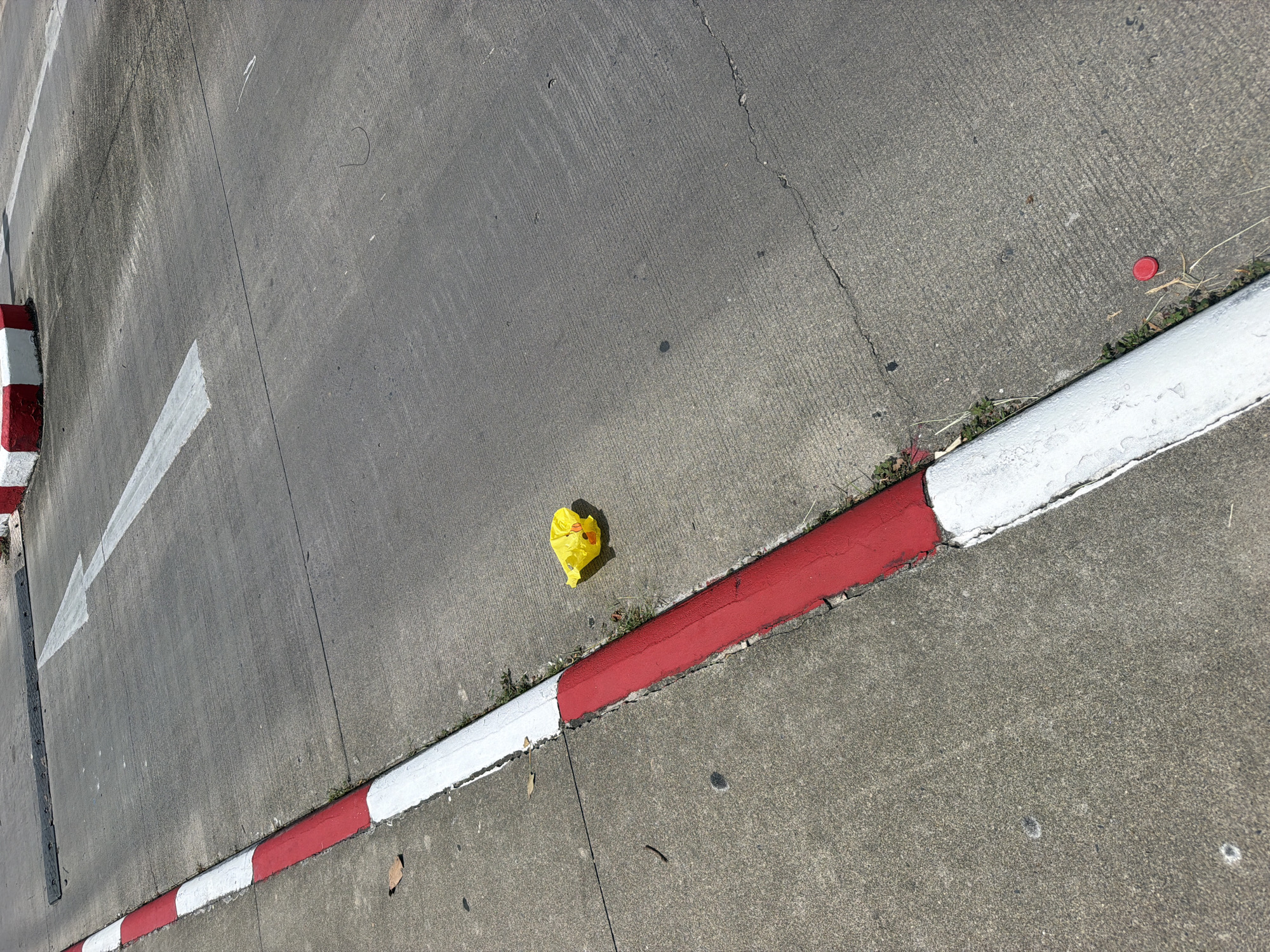 The image depicts a concrete road in Ko Samui, Thailand, marked with red and white striped curbs. A crumpled yellow plastic bag lies on the road near the curb. White arrows are painted on the road surface, indicating direction. In the background, a red and white concrete barrier is visible. The scene appears to be a public area, possibly a street or parking lot.