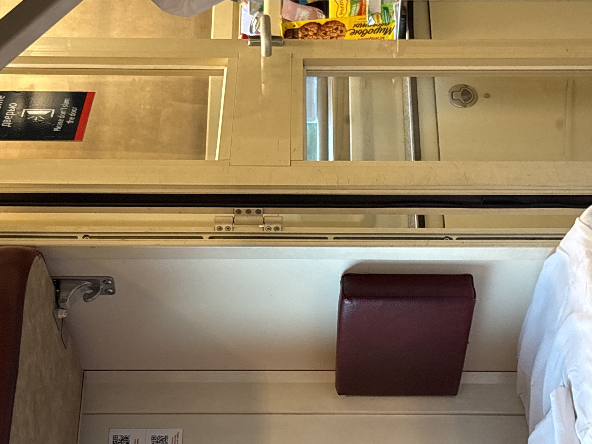 This media file shows an interior view of a train compartment. The scene is viewed from a low angle, looking upwards. The subjects are primarily objects within the compartment, including shelving, a small dark red seat or cushion, and a white, rumpled fabric, likely bedding or clothing, on the right.
On the upper shelf, there are several items, including a yellow and blue packaged snack or food item with text in Cyrillic. Next to it, partially obscured, is another item with similar packaging.
In the middle section, a black and red sign is visible on the left, attached to a light-colored surface. The sign contains text in both Cyrillic and English. The English text reads "Please don't slam the door." The Cyrillic text is partially visible and appears to be related to the door.
Below this, a dark red, upholstered object, possibly a seat or a bolster cushion, is positioned on the right. To the left of it, a metal bracket is attached to a light-colored wall.
The overall setting suggests a train compartment, possibly a sleeping berth, given the presence of the bedding-like fabric and the shelving for storing items. The lighting appears to be artificial or ambient, and there are no clear indicators of the time of day or weather. The location context provided is Shchekutovo, Russia. No people are visible in the frame. The visible text provides a clear instruction for passengers.