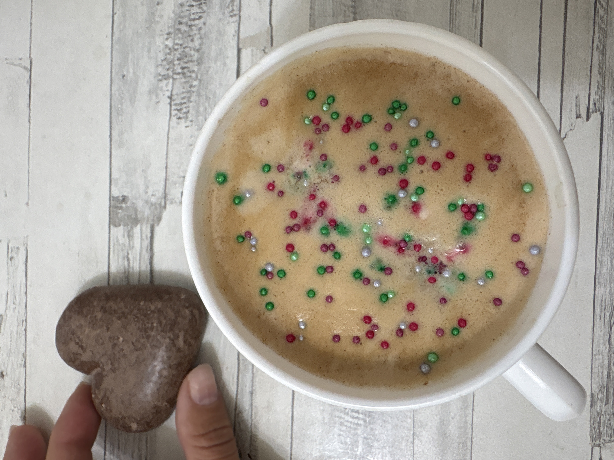 A top-down shot features a white mug filled with a light brown, frothy beverage speckled with red, green, and silver sprinkles. To the left of the mug, a hand holds a heart-shaped brown cookie. The background consists of a light grey wood-grain surface. The scene appears to be a coffee break with a festive touch. No location in the city of Chaikovsky, Russia is discernable.