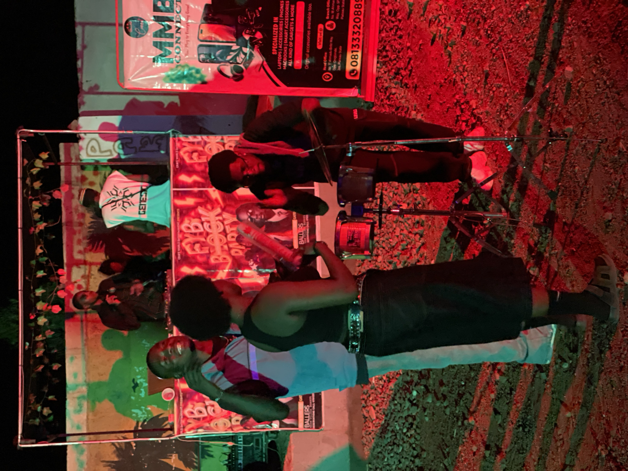An outdoor nighttime scene in Durumi, Nigeria, depicts a gathering with music and what appears to be a promotional event. In the foreground, a person wearing a black tank top and a dark skirt is playing a drum set. To their left, another individual in a striped shirt is looking towards the drum player. In the background, on a raised platform, a person in a black jacket is speaking into a microphone, and another individual in a white t-shirt with "GENESIS" printed on it is visible. A large banner to the right advertises "MMB CONNECT" and specializes in electronics. The ground is covered in gravel and illuminated by red and green lights.