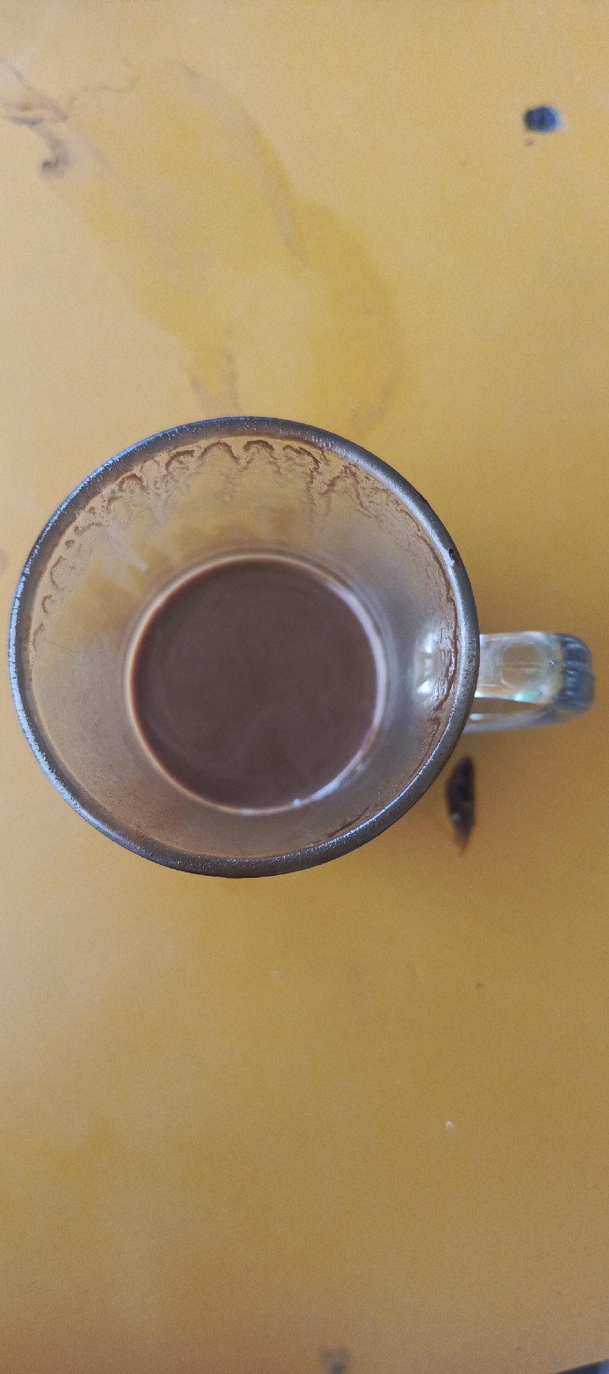 A clear glass mug containing a brown beverage, likely coffee or tea, rests on a yellow, textured surface. The mug has a handle and is partially filled, with a visible residue line on the inside of the glass near the rim. The surface appears to be a table or countertop, with some slight variations in color and texture. There are no people visible in the image. The lighting suggests an indoor or shaded outdoor setting. The image is a top-down view of the mug and its immediate surroundings. The location is Kletek, Indonesia.