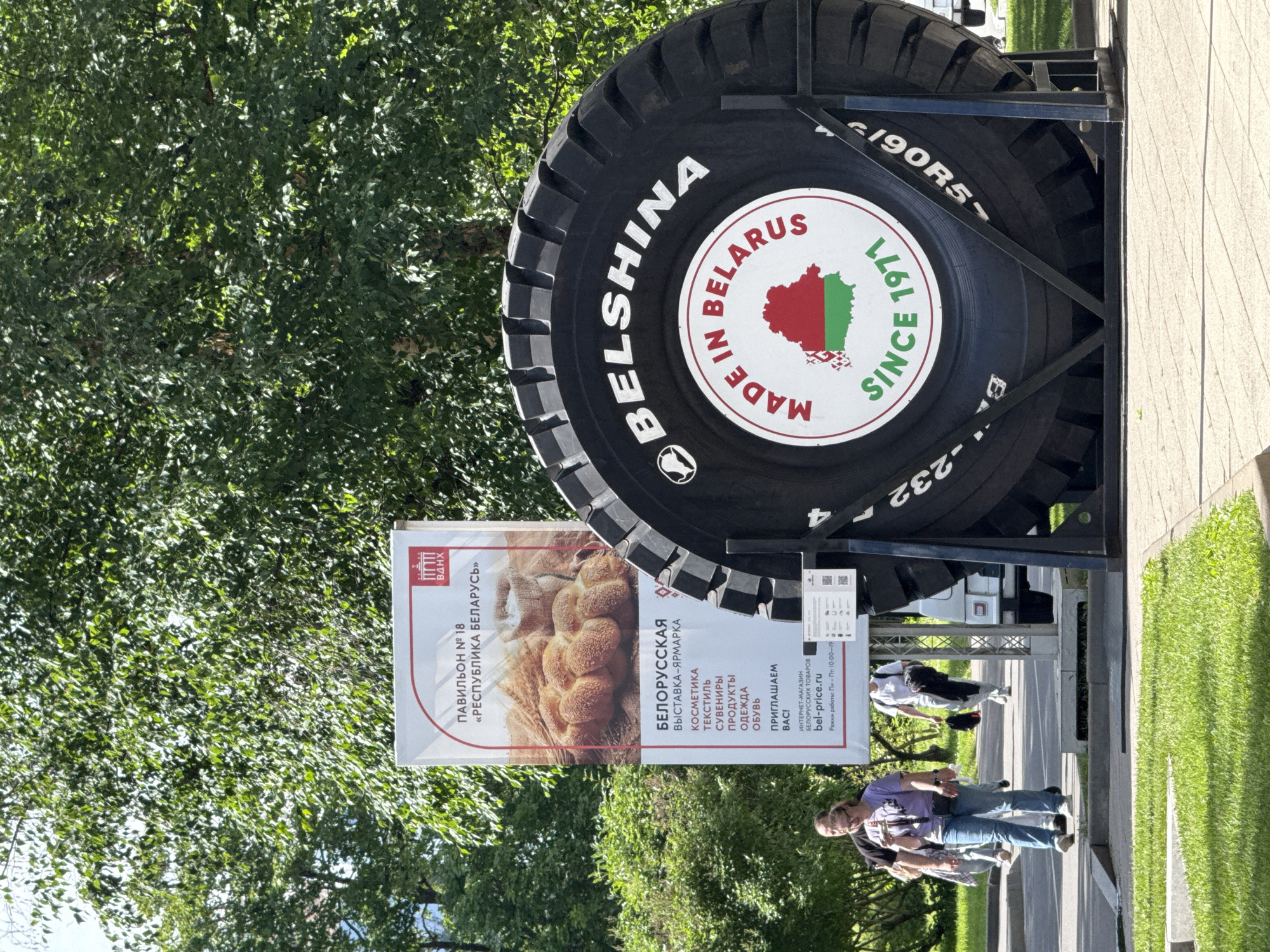 This media file captures an outdoor scene in Moscow, Russia, on a bright, sunny day. The primary focus is a giant tire, likely a promotional display, with "BELSHINA" and "MADE IN BELARUS SINCE 1971" prominently visible on its surface, along with the Belarusian flag within a map outline. The tire is mounted on a black metal stand, with another tire visible behind it.
In the background, lush green trees provide shade. To the left of the tire, a large banner advertises a "BELORUSSKAYA VISTAVKA-YARMАРКА" (Belarusian Exhibition-Fair) in Pavilion No. 18, "REPUBLIKA BELARUS". The banner lists various Belarusian goods for sale, including cosmetics, textiles, souvenirs, food products, clothing, and footwear, and provides a website and operating hours for an online store.
Several people are present in the scene, mostly walking along a paved area in front of the banner and tire display. One person in the foreground is wearing a purple t-shirt and blue jeans, while another person in the mid-ground is wearing a white t-shirt. The overall atmosphere appears casual and commercial, suggesting a public event or market. The presence of the tire and the exhibition banner indicates a promotion of Belarusian products.