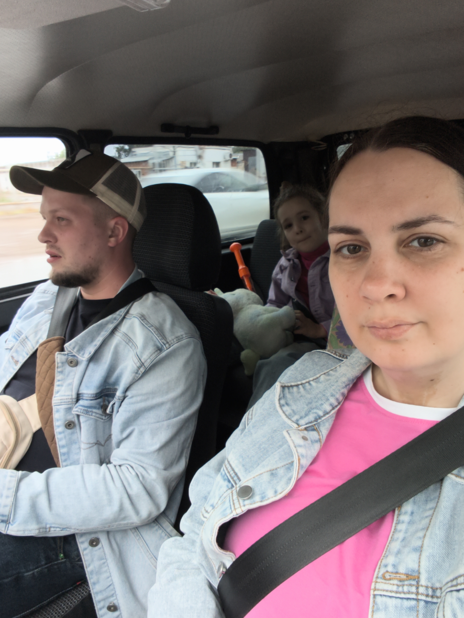 The image captures three individuals inside a car. In the front passenger seat, a woman with dark hair is wearing a pink shirt and a denim jacket. She is wearing a seatbelt and appears to be taking the photograph. To her left, in the driver's seat, is a man with short hair and a beard, wearing a baseball cap, a denim jacket, and a black shirt. He is also wearing a seatbelt. In the back seat, a young girl is seated, wearing a purple shirt and jacket. There's an orange plastic toy and a stuffed animal next to her. The car's interior is visible, including the headrest of the driver's seat. Through the windows, a blurry street scene with a white car and buildings suggests an urban environment. Based on the prompt mentioning Chaikovsky, Russia, it is assumed to be the location.