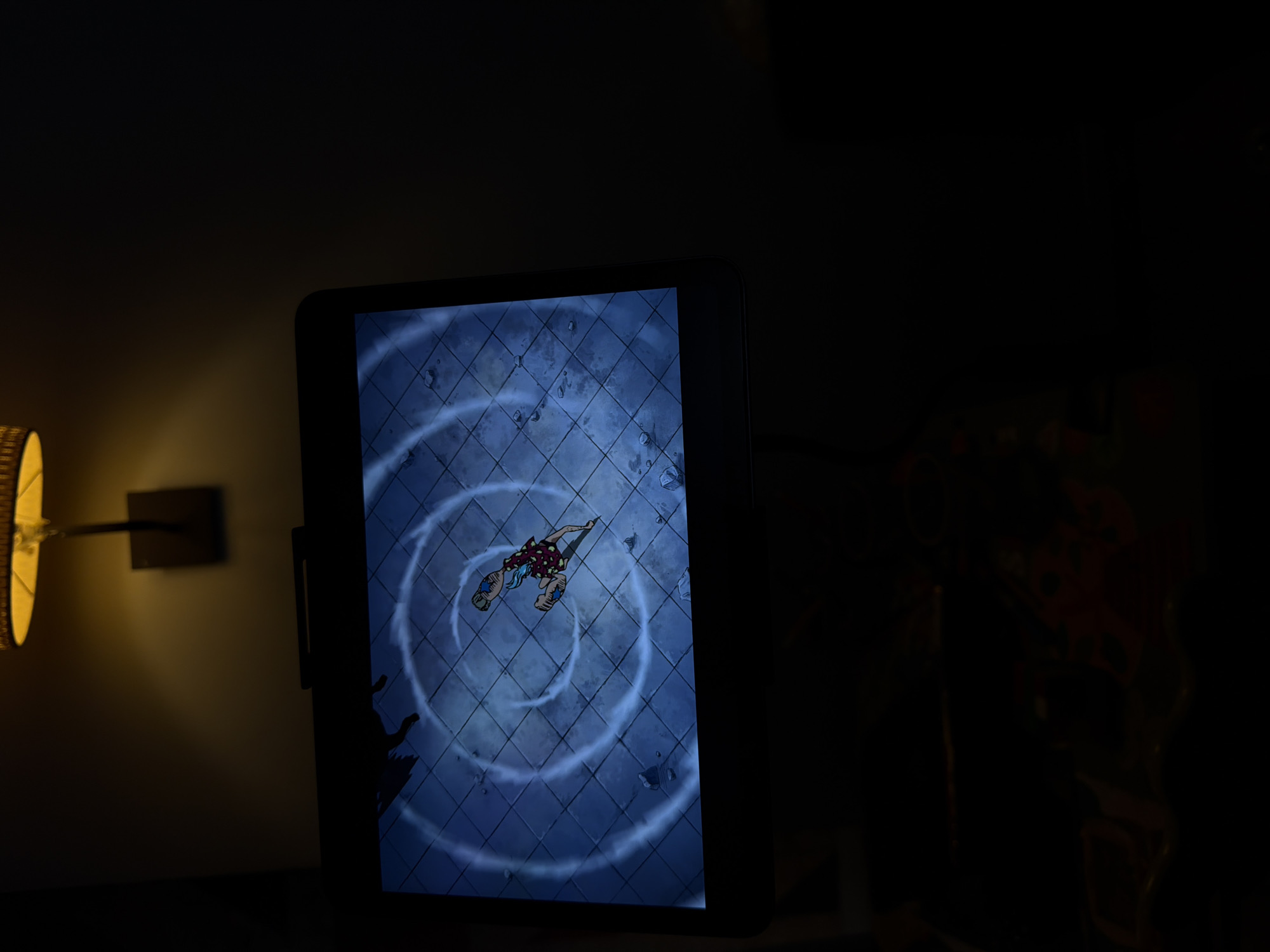 A dimly lit room displays a tablet or screen showing a digital image. The screen depicts a cartoonish figure floating or falling in a circular pattern over a tiled blue-grey surface. A lamp with a wicker shade is visible on the left. The background is very dark, with an indistinct object possibly a plant. There is no indication of the location being Sofia, Bulgaria, or any specific city.