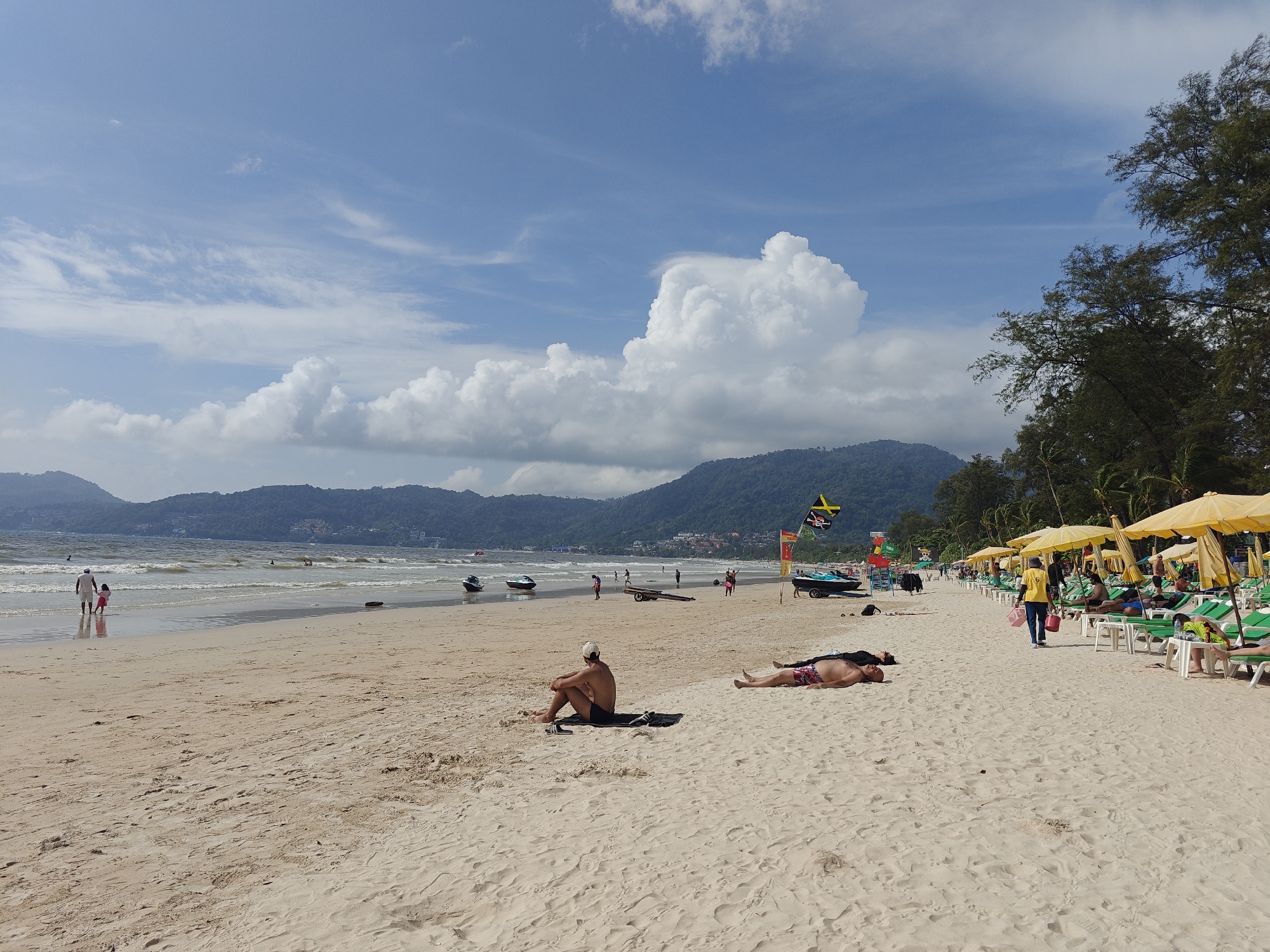 The image depicts a sunny day at Patong Beach, Thailand, a popular tourist destination. The foreground is dominated by fine, light-colored sand, marked with footprints and the gentle undulations of the tide. In the middle ground, several people are visible. Two men are lying on towels on the sand, sunbathing. One man is sitting upright, facing away from the camera, with his legs crossed. Further down the beach, closer to the water, a man and a child are walking by the shoreline. The sea is calm with small waves, and several jet skis are parked on the sand. In the background, lush green hills rise behind a line of buildings and palm trees, characteristic of the Patong coastline. A row of yellow beach umbrellas and green lounge chairs is set up along the edge of the sand, indicating a developed beach resort area. The sky is a vibrant blue, dotted with fluffy white clouds, some of which are quite large and dramatic. Flags, including one with a Jamaican motif, are visible near the beach umbrellas. The overall scene is one of a relaxed tropical beach environment.