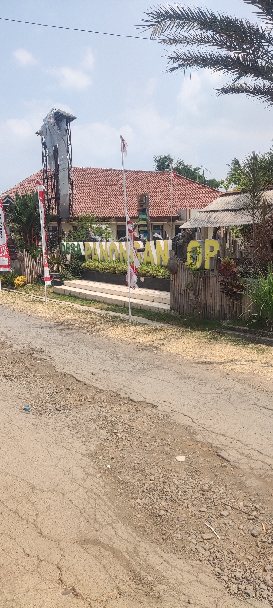 In Panongan Lor, Indonesia, a sign reading "DESA PANONGAN" is prominently displayed with large, yellow, three-dimensional letters. To the right of this sign, the letters "OP" are also visible, also in large, yellow, three-dimensional letters. The sign is situated in front of a building with a red tiled roof. Several Indonesian flags, featuring red and white stripes, are visible, some are mounted on poles. A tall structure, appearing to be a tower with a weathered, metallic covering and some dangling materials, stands to the left of the sign. The area in front of the sign features a short set of stairs leading up to a paved area. The foreground of the image shows a cracked asphalt road with patches of gravel and dry grass. Palm fronds are visible at the top of the frame, suggesting a tropical environment. The sky is bright and appears to have some clouds.