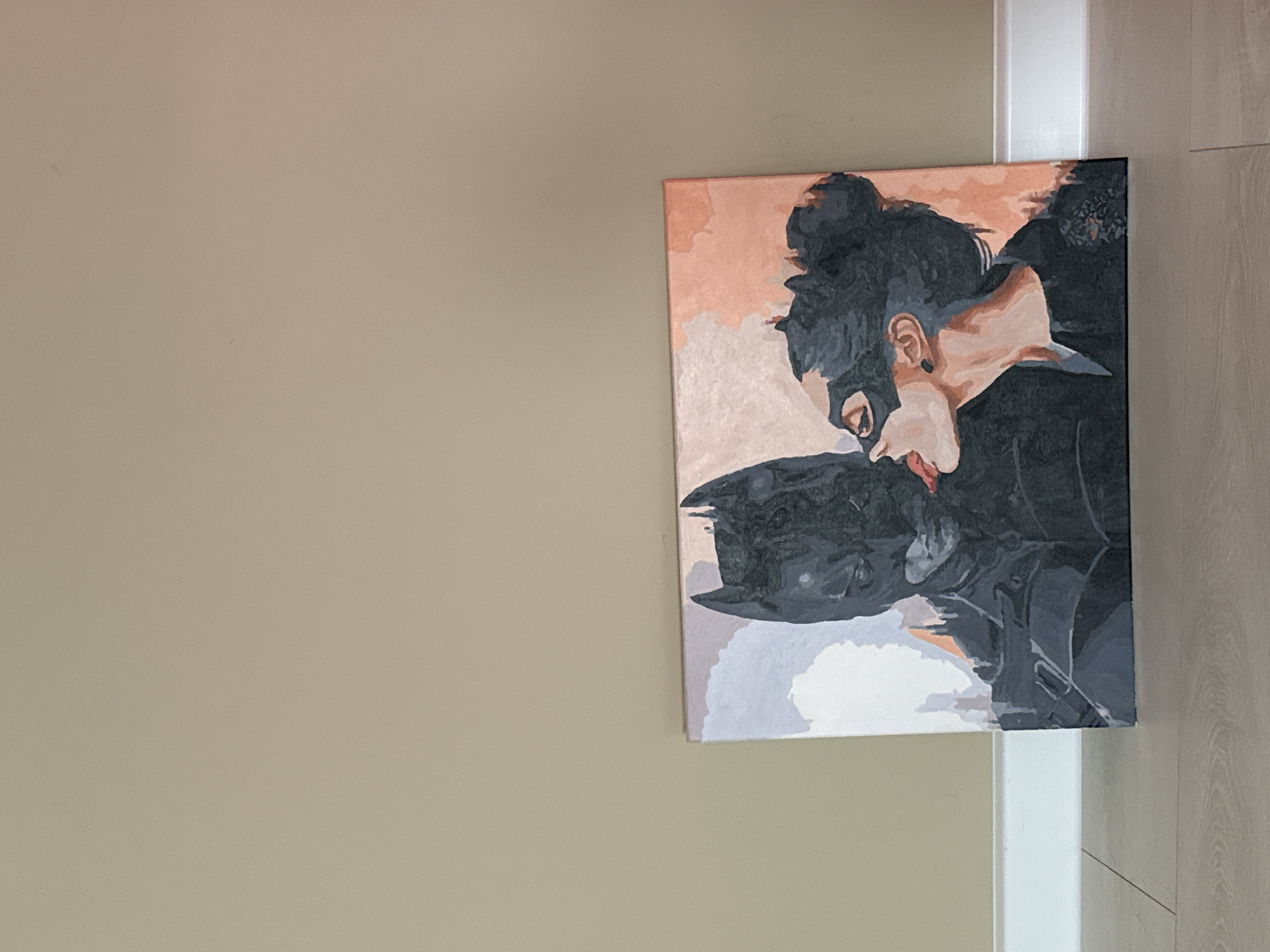 A painting of Batman and Catwoman hangs on a beige wall next to a white door frame and wood-paneled wall. The painting depicts Catwoman in a dark gray costume with her hair in a messy bun, leaning over Batman, whose hat is visible. The background of the painting is a mix of peach and light gray.