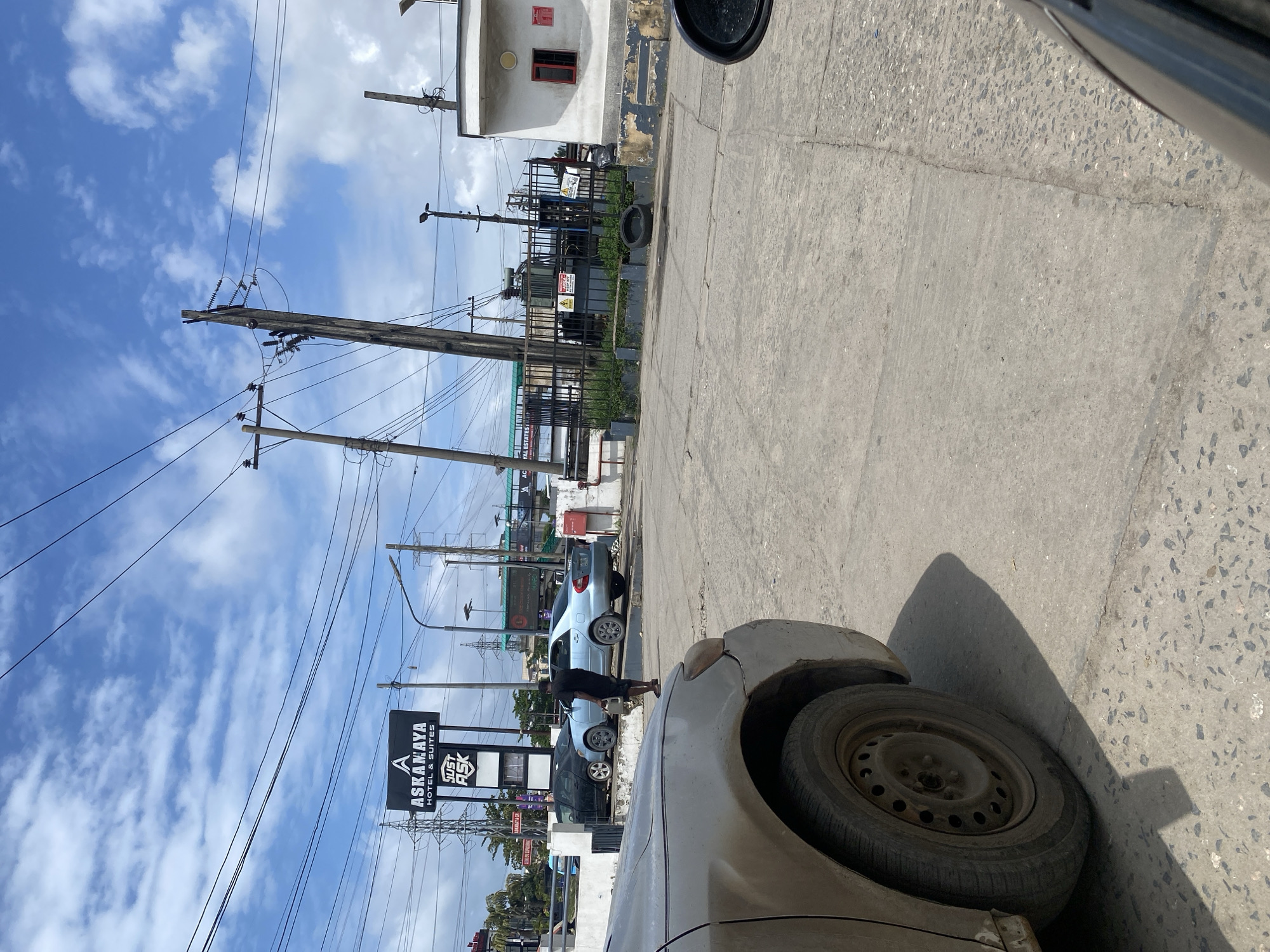 A man carrying a white jerrycan walks across a paved road in Ajah, Nigeria, towards a light blue sedan parked parallel to a black sedan. In the background, a prominent sign reads "ASKANAMAYA HOTEL & SUITES" with smaller text below that says "JUST ASK". Further back, there is an electrical transformer station enclosed by a black metal fence, a small white building with a red-framed window, and various electrical poles and wires against a cloudy blue sky. The foreground is dominated by the front passenger side of a dirty, light-colored car.
