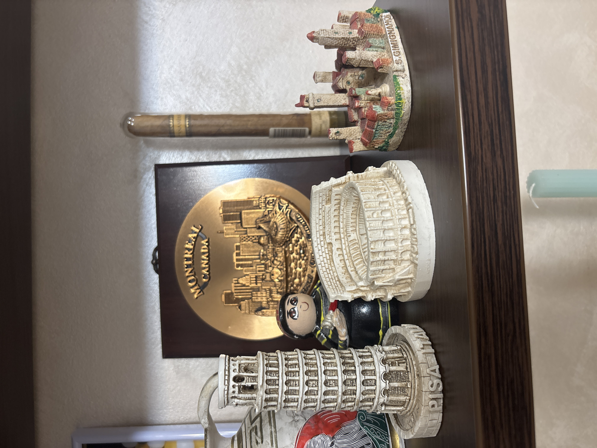 The image shows a collection of souvenirs displayed on a shelf against a light-colored wall. The souvenirs are arranged in a vertical row, primarily representing cities and landmarks. Starting from the top, there is a cigar in a plastic tube, followed by a souvenir of San Gimignano, a gold-colored Montreal souvenir, a souvenir of the Colosseum, a souvenir figure, and a souvenir of the Leaning Tower of Pisa. The shelf has a dark brown wooden frame.
There is no information in the image to suggest that this scene is in Plovdiv, Bulgaria.