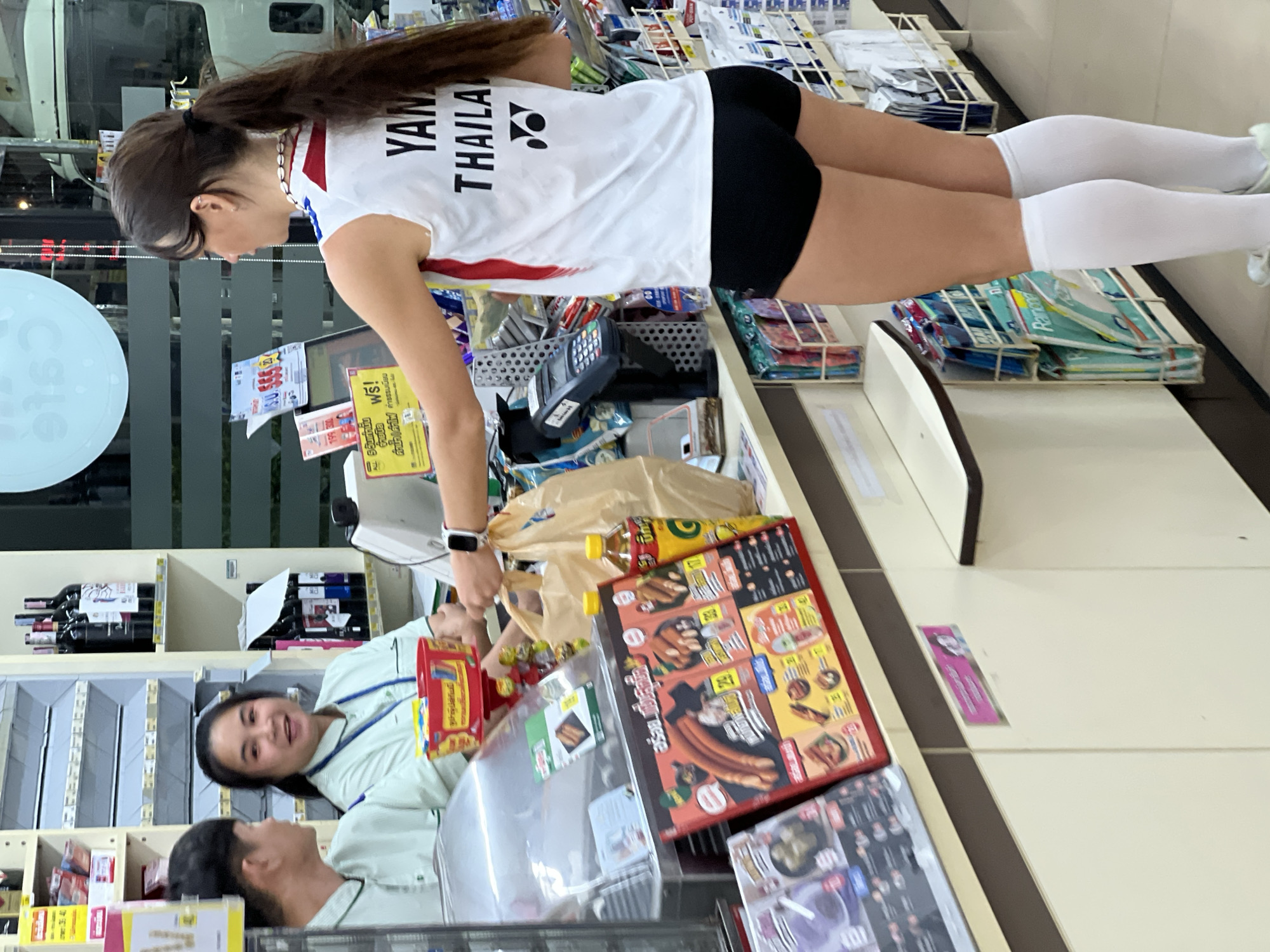 The image depicts a scene inside a convenience store in บ้านบางรักษ์, Thailand. A fair-skinned woman in a white shirt with "YAN THAILA" printed on the back and black shorts, along with white socks, is at the checkout counter. She's handing a shopping bag to the store clerk, a light-skinned woman with short black hair wearing a light green uniform. Various products, including packaged foods and beverages, are visible on the checkout counter and shelves behind the clerk. A sign and a credit card machine are positioned between the customer and the clerk.