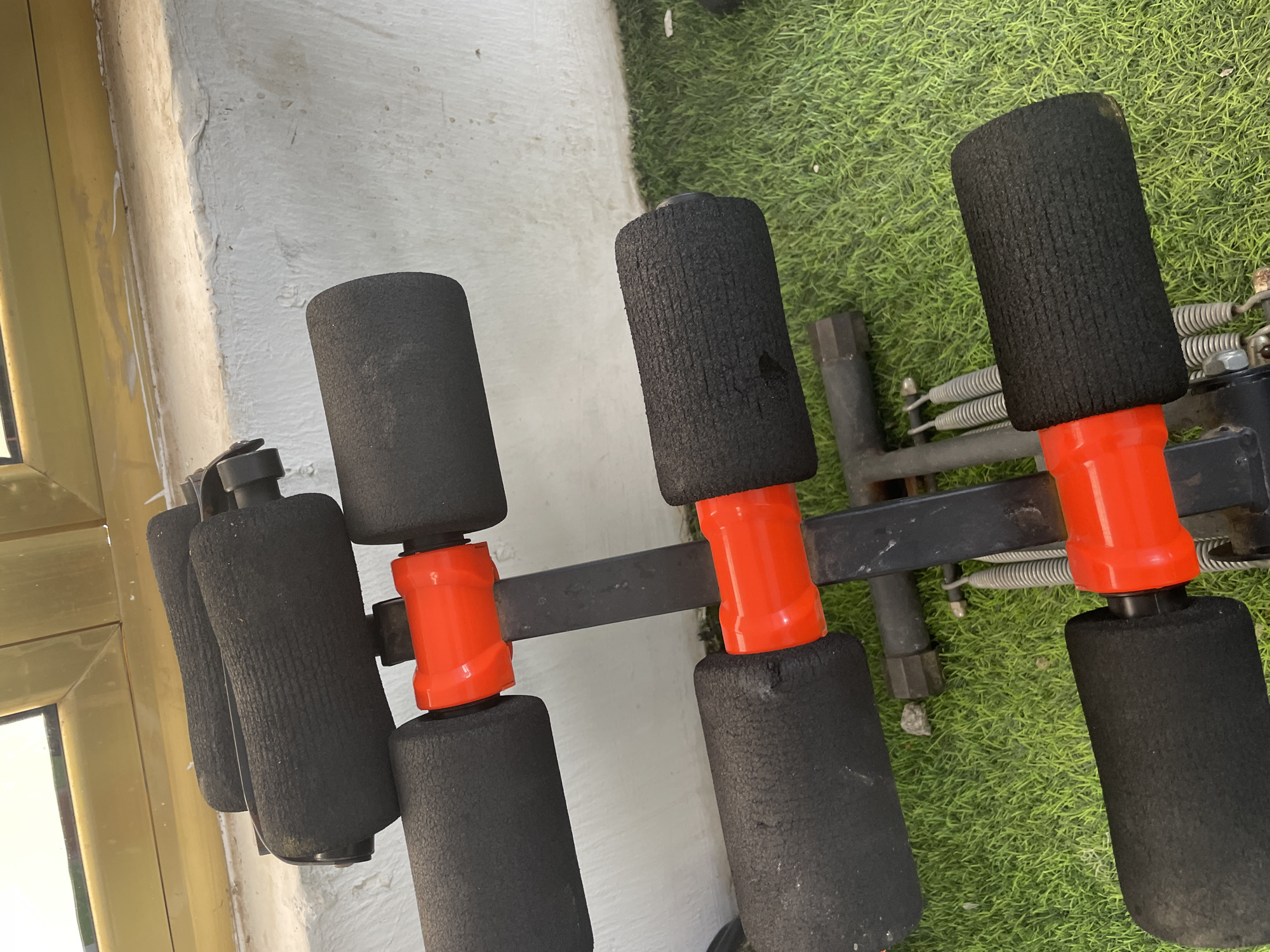 A close-up, high-angle shot shows a black and orange exercise bench with foam rollers. The bench is positioned on a patch of artificial green grass, likely located outdoors. In the background, a white textured wall meets a yellow-painted window frame. The scene appears to be in the city of Jauro Yinu, Nigeria. There are no people or animals visible in the frame.