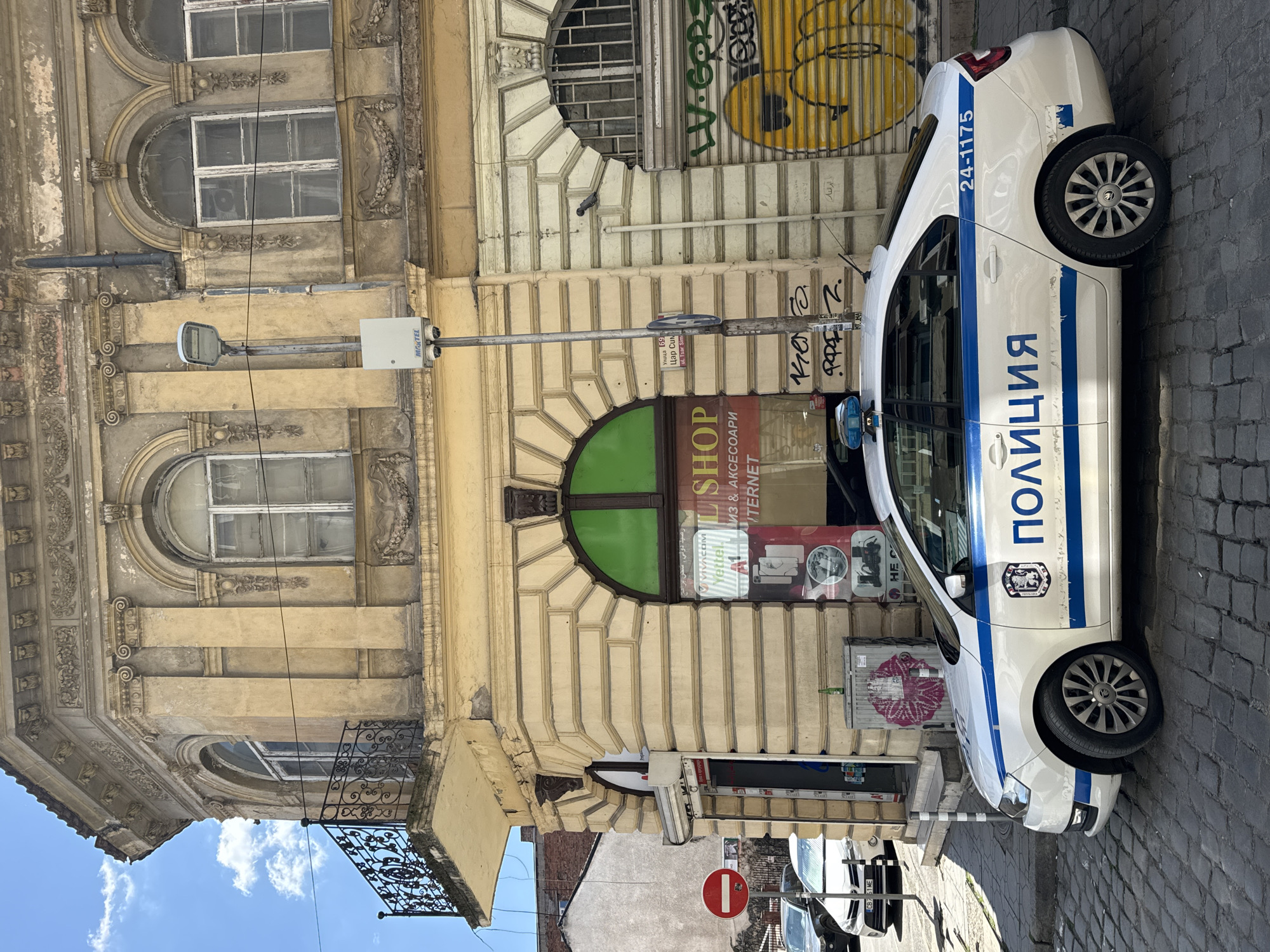 A white police car with blue stripes is parked on a cobblestone street in Sofia, Bulgaria, next to an ornate, multi-story building. The vehicle has "ПОЛИЦИЯ" (POLICE) written in blue Cyrillic letters along its side, along with the number "24-1175" and a national crest. The building features arched windows, decorative architectural elements, and worn light-colored stucco.
On the ground floor of the building, a shop is visible with a green-framed arched entrance and window displays. Signs on the shop read "SHOP", "МИЗ & АКСЕСОАРИ" (Mice & Accessories), and "ИНТЕРНЕТ" (Internet). Further text includes "VIVACOM" and "Yettel" (Bulgarian telecom providers), and some Arabic script: "رصيد دولي, سحن برامج" (international balance, program charging) and "الد العالم" (World of...). Graffiti in yellow and green is present on a metal shutter above the shop.
A street lamp post stands near the police car, featuring a light fixture and a white box with "MAKTEL" written on it. A small sign attached to the post reads "Улица 69 Цар Сим" and "ul. Tsar Sim" (referring to Tsar Simeon Street). Another red circular "no entry" sign is visible further down the street. The sky is bright blue with scattered white clouds, indicating daytime with clear weather. Partial views of other parked cars and architectural elements like an iron balcony are also present.