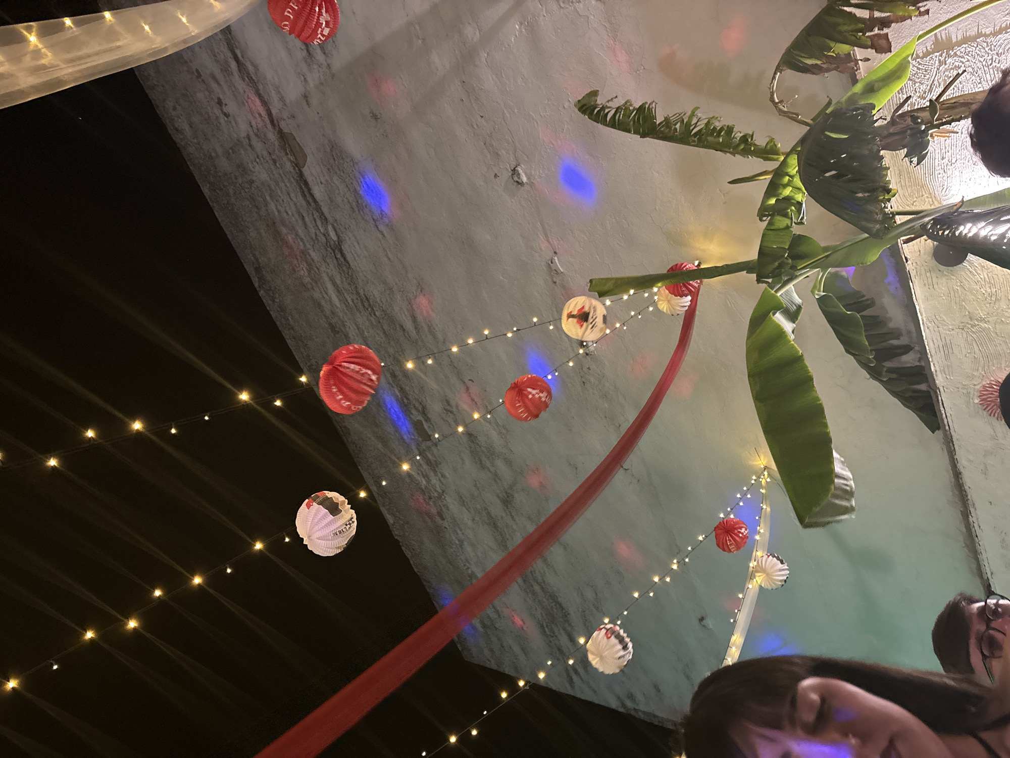 The image depicts an outdoor event, possibly a party or gathering, in Córdoba, Spain. The location appears to be an open courtyard or terrace, decorated with festive elements like string lights, paper lanterns in red and white, and a red fabric banner draping across the ceiling. Tropical foliage, potentially banana plants, adds a vibrant touch to the setting. The background reveals a textured, light-colored wall illuminated by colorful lighting effects (blue, pink). Parts of individuals are visible, including faces and hair, suggesting a social gathering. The overall impression is of a celebratory atmosphere.