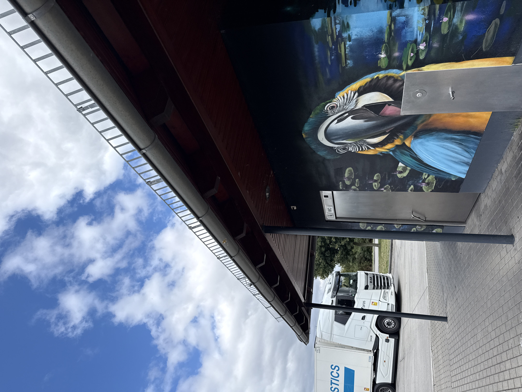 The image depicts an outdoor scene under a partly cloudy sky. In the foreground, a paved area with cobblestones leads to a building with a mural of a blue and yellow macaw. A metal door with a "WC" sign is visible on the building. A metal water fountain is mounted on the mural. To the left, a white DAF semi-truck is parked. The truck has "LOGISTICS" written on its side. In the background, trees and a cloudy sky are visible. The location is identified as Großpösna, Germany.
