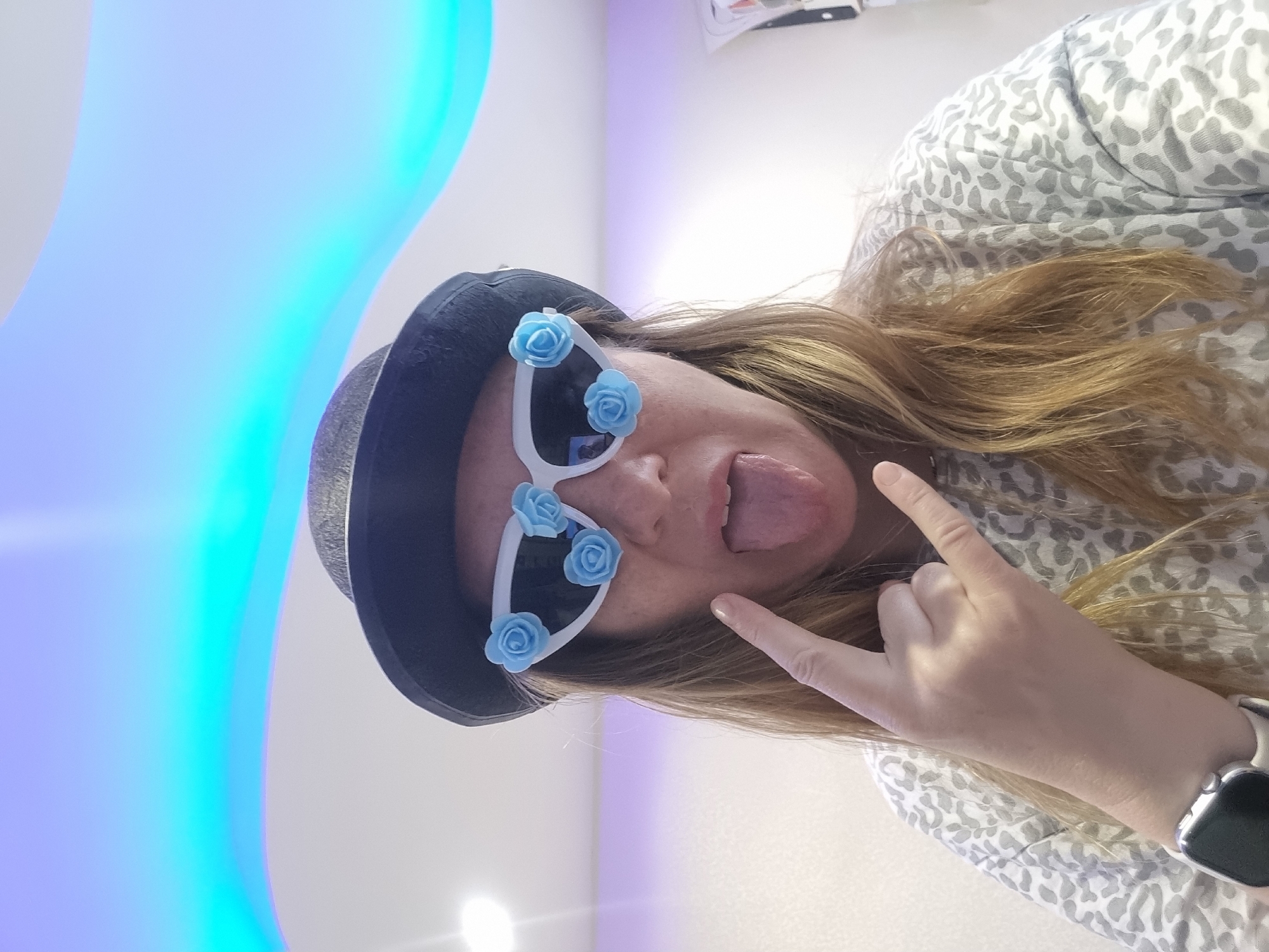 An adult female is captured in a close-up, angled shot, displaying a playful and energetic expression. She has long, light brown hair and visible freckles on her face. She is wearing a dark, possibly black, fedora-style hat. Unique white sunglasses, each decorated with three light blue artificial roses on the frames, cover her eyes. Her tongue is playfully sticking out, conveying a mischievous mood.
Her left hand is raised, forming the "rock and roll" or "devil horns" gesture, with the index finger and pinky extended. She is wearing a light grey and white patterned long-sleeved shirt, featuring an animal print design. A smartwatch with a silver-toned body and a white band is visible on her left wrist.
The background consists of a smooth, light-colored wall, illuminated by a vibrant, wavy light feature that transitions through shades of blue, cyan, and purple. This artificial lighting suggests an indoor setting, possibly an entertainment venue or a specially decorated room in Chaikovsky, Russia. There is no visible text in the image.