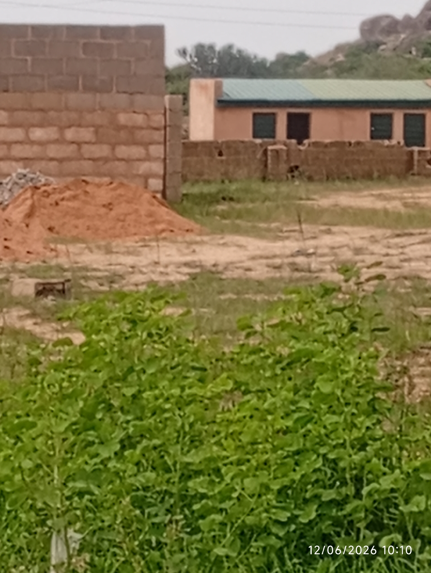 The image depicts a construction site in Du, Nigeria, with a partially constructed brick wall on the left and a building with a green roof in the background. A large pile of sand is visible in the foreground. Lush green vegetation covers the lower portion of the image, with a date and time stamp "12/06/2026 10:10" in the bottom right corner. The overall scene suggests an ongoing development project in a semi-arid environment, with rocky hills in the far background.