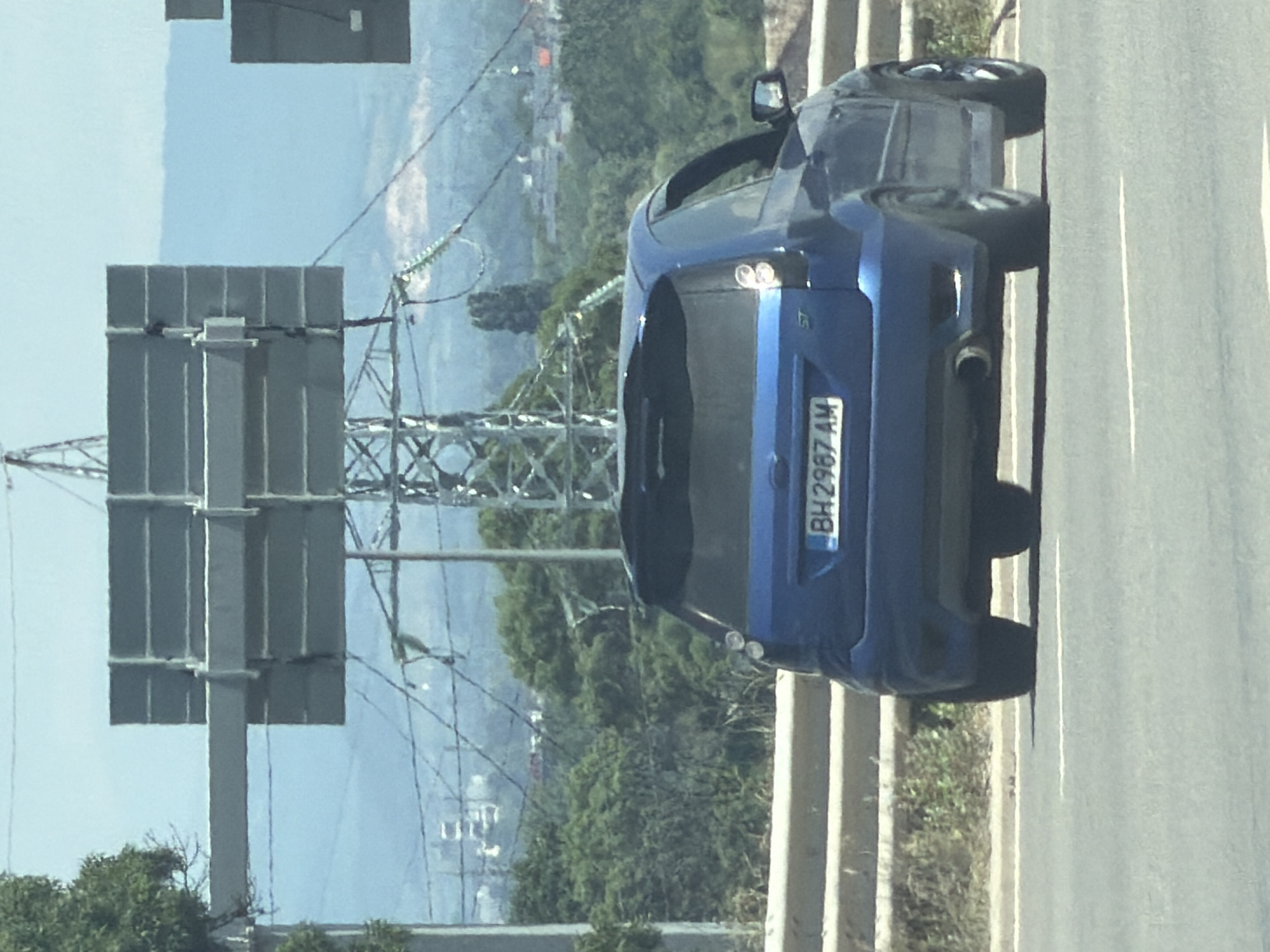 A blue Ford Fiesta ST is driving on a road in Sofia, Bulgaria. The car is seen from the rear, and the license plate reads "BH 2987 AM". The car is in the right lane of a two-lane road, with a concrete barrier to its left. In the background, there are trees, power lines, and a large metal structure with rectangular panels, likely a billboard or an informational sign. In the distance, there are hills and a hazy sky. The overall environment appears to be semi-urban or suburban, with a mix of infrastructure and natural elements. The lighting suggests it is daytime, with bright sunlight.