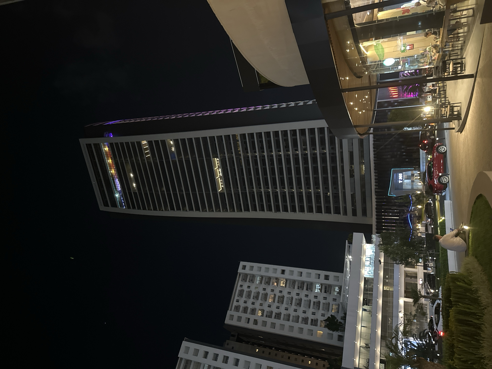 This nighttime photograph depicts a cityscape of Heroica Puebla de Zaragoza, Mexico. A tall, slender skyscraper, illuminated with rows of horizontal lights, stands out against the dark sky. Below the skyscraper, there are two lower-rise white buildings. The foreground shows the exterior of a restaurant or cafe, with visible tables and chairs inside. A red car is parked on the right side of the frame, along with other parked cars. A person is sitting on the edge of a planter box, which is bordered by green plants. The overall scene is brightly lit by artificial lights.