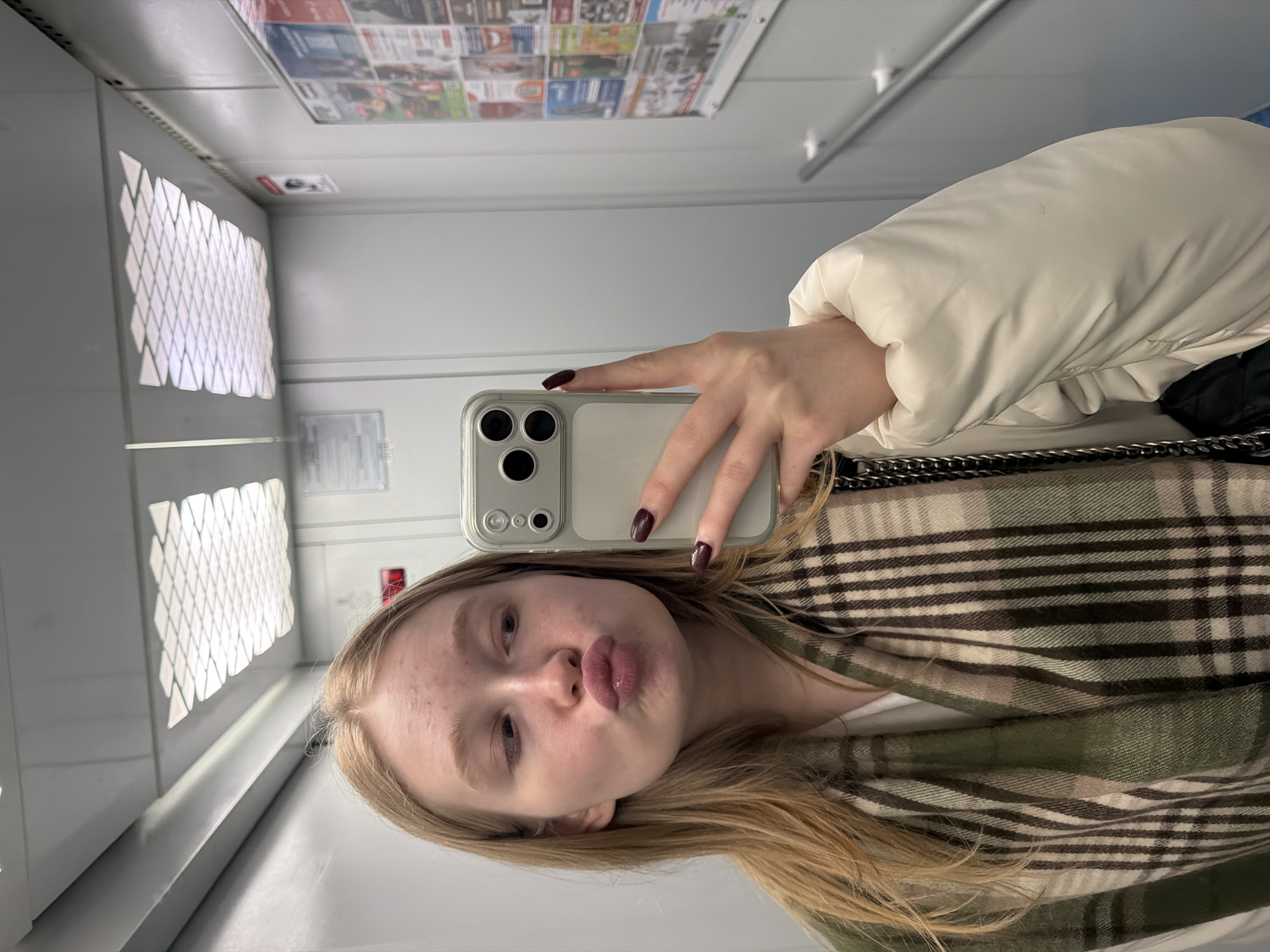 A young woman is depicted taking a mirror selfie in an indoor setting, possibly an elevator or hallway, located in Penza, Russia. She has long, straight blonde hair and fair skin, and is making a "duck face" expression towards the camera. Her fingernails are painted dark maroon. She is wearing a cream-colored puffer jacket and a green and beige plaid scarf or shawl.
The woman holds a light-colored smartphone, appearing to be an iPhone with a triple camera setup, encased in a clear protective cover. The selfie is captured via a reflective surface, which shows the environment upside down due to the image's orientation.
The background consists of light-colored, highly reflective walls and a ceiling. On the left, a prominent light fixture with a diamond or geometric pattern illuminates the space. The ceiling is covered with numerous small papers or advertisements, though their text is illegible due to distance and reflection. On the right wall, a faint white rectangular sign with unreadable text and a red object, possibly an emergency button or fire alarm, are visible. The overall impression is of a brightly lit, modern interior space.