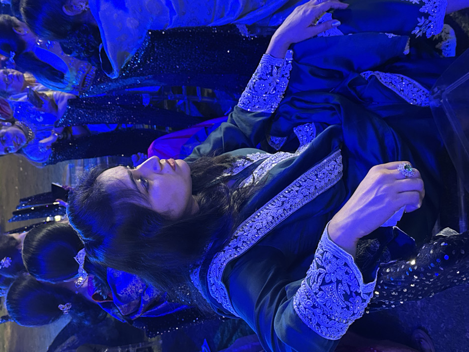 A woman in a deep blue, ornate outfit reclines among a group of similarly dressed women. Her dress features intricate silver embroidery along the neckline and sleeves. She wears a large emerald ring on her right hand, which is held aloft. Her dark hair is partially pulled back, and she gazes upwards with a serene expression. The lighting creates a blue, theatrical atmosphere. Other women in sparkling dresses are visible in the background, suggesting a formal event or performance.