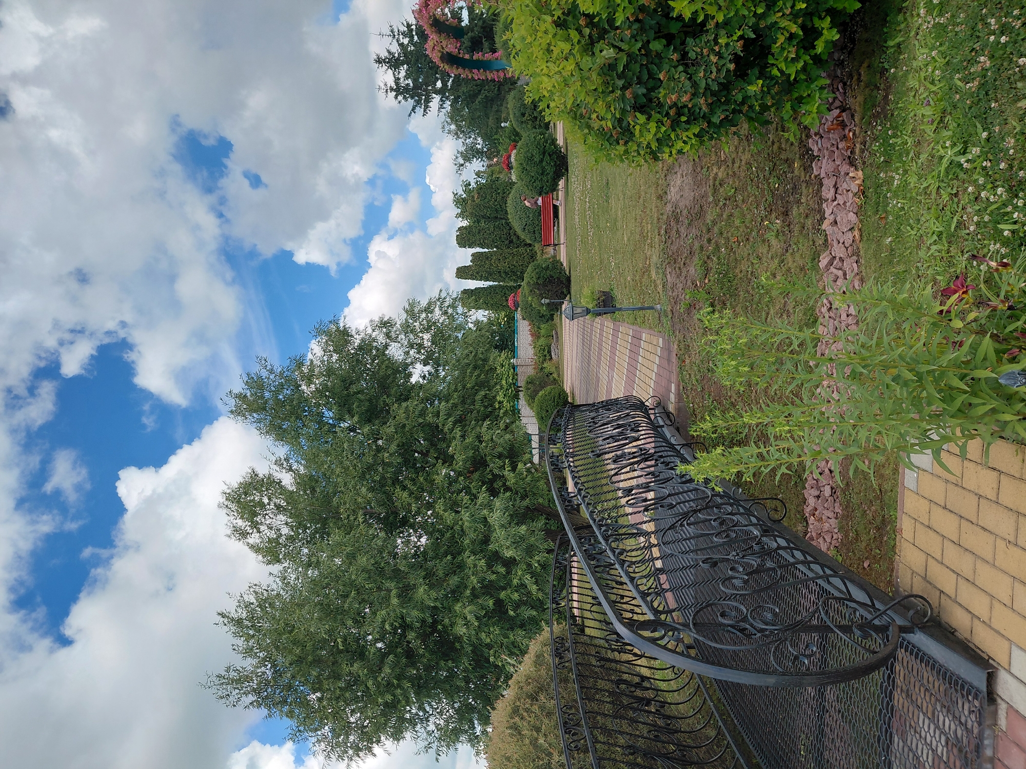 The image depicts an outdoor scene in the city of Kovalivka, Ukraine, likely a park or garden area. In the foreground, a decorative black metal bridge with intricate scrollwork spans across a paved walkway made of yellow and red rectangular bricks. To the left of the bridge, lush green foliage is visible. Beyond the bridge, a neatly manicured lawn is dotted with various shrubs and trees. In the middle ground, a person is seated on a red bench situated on the grass. Further in the background, more trees and a decorative floral archway can be seen against a bright blue sky filled with fluffy white clouds. The overall atmosphere suggests a pleasant, well-maintained outdoor recreational space.