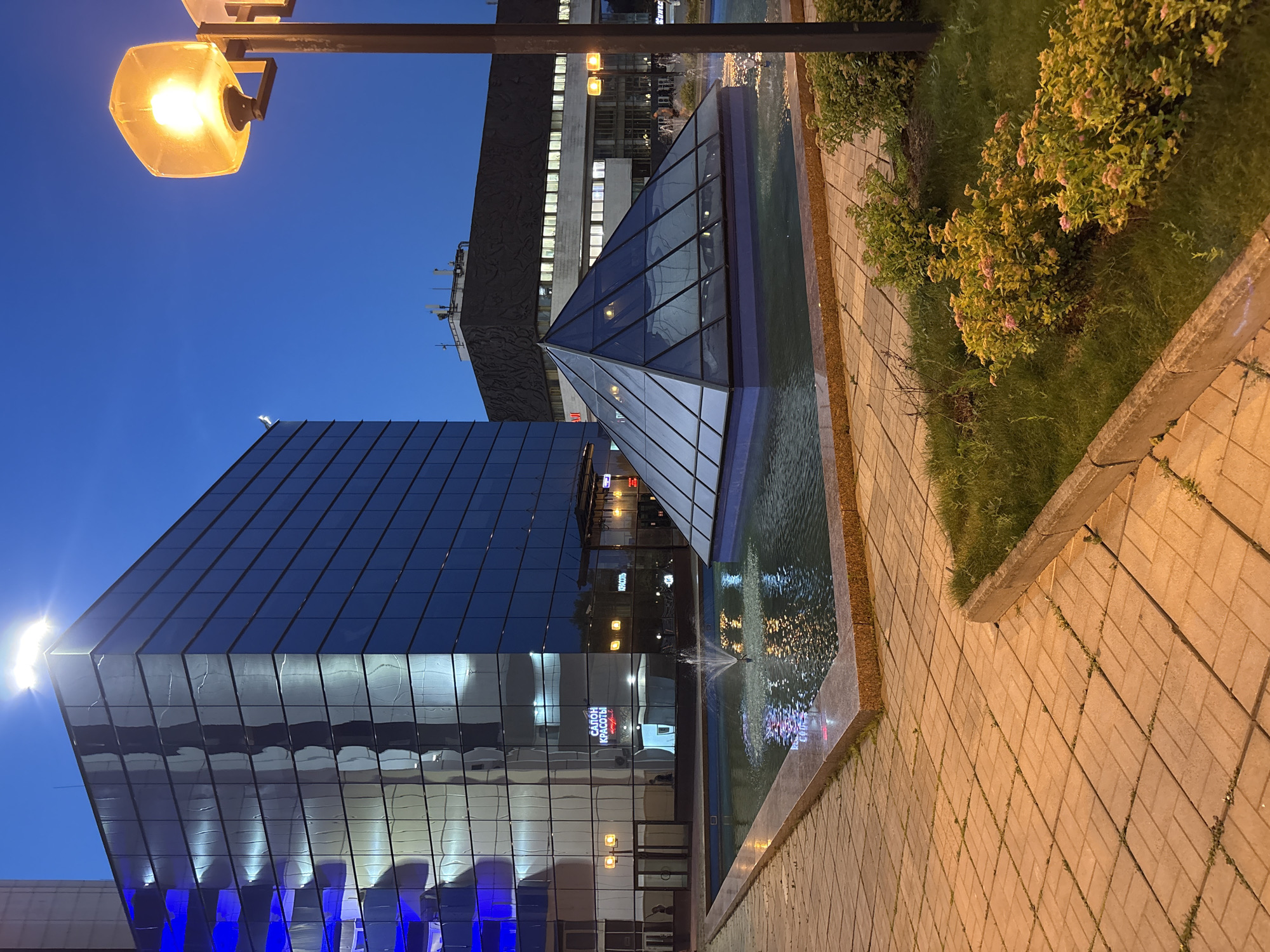The image depicts a street scene in Moscow, Russia, during twilight. Dominating the left side is a modern, glass-clad building with blue lighting illuminating its upper floors. In front of this building, a shallow, rectangular fountain features a glass pyramid structure in its center, with water jets creating a spray. The fountain is surrounded by a paved walkway made of light-colored rectangular bricks. To the right of the fountain and building, a second, larger building with a textured dark facade and illuminated windows is visible. A lamppost with a bright, warm-toned light stands in the foreground on the right. Lush green grass and flowering bushes are present in a landscaped area to the right of the paved walkway. The sky is a deep blue, indicating the transition from day to night. A sign with Cyrillic lettering, "САЛОН КРАСОТЫ," is visible on the glass building, translating to "Beauty Salon." Several indistinct figures can be seen through the glass doors of the building and in the background.