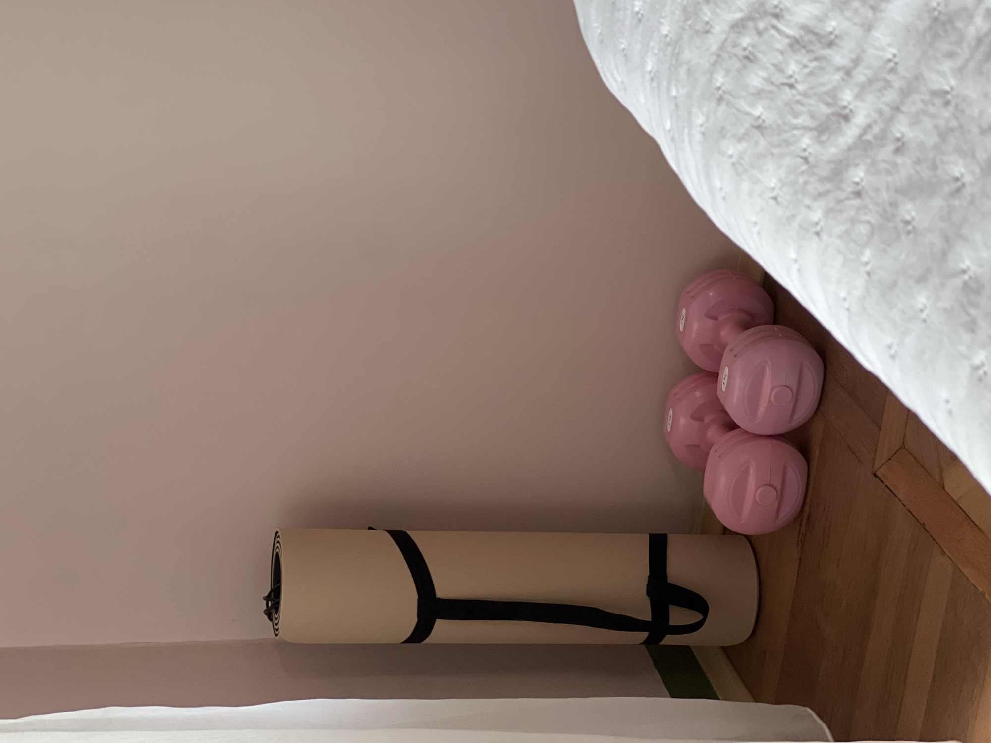 In the corner of a room, a beige yoga mat is rolled and secured with black straps, standing upright on a polished wooden floor. To the right of the yoga mat, three pink, spherical dumbbells are placed on the floor. A white, textured comforter partially covers the right side of the frame, suggesting the presence of a bed. The walls are painted a soft pink color, and a light-colored curtain hangs on the left side of the image. The scene is indoors, with no indication of the city of Sinyavino, Russia, or any external environment.
