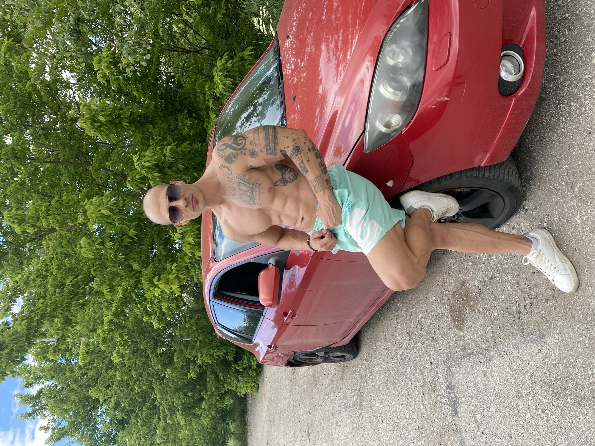 A shirtless, muscular, bald man wearing sunglasses and light blue shorts poses with a red Mazda automobile, with his left foot resting on the car's front tire. The car is parked on an asphalt surface, with green foliage visible in the background. The man's body features numerous tattoos. The location may be in Pleven, Bulgaria, but this cannot be definitively confirmed from visual evidence.