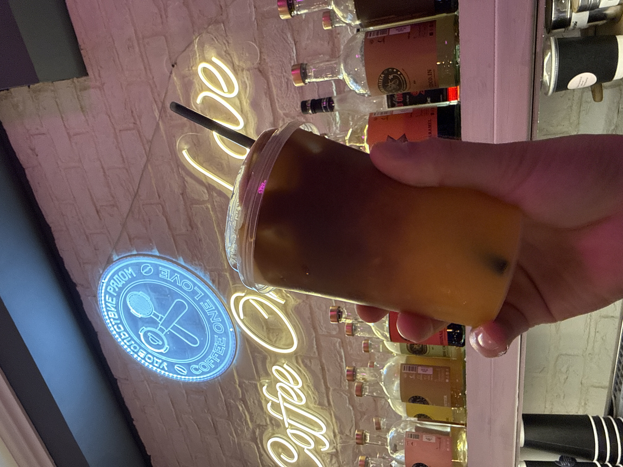 The image displays a close-up, tilted view of a person's hand holding a layered iced beverage in a clear plastic cup with a black straw. The drink consists of a dark brown top layer, likely coffee, and a lighter, possibly orange-colored, bottom layer.
The background features a brick wall adorned with neon signs. A yellow neon sign, partially visible, spells out "Coffee O love". Below and to its left is a prominent circular blue neon sign. This blue sign showcases a graphic of a coffee portafilter and tamper, encircled by text in both Cyrillic and English. Visible phrases include "ONE LOVE COFFEE," "СЧАСТЬЕ РЯДОМ" (Happiness is near), and "УДОВОЛЬСТВИЕ" (Pleasure/Enjoyment), which aligns with the given location context of Khimki, Russia.
Behind the signs and the hand, shelves hold numerous glass bottles, possibly containing syrups or spirits, along with stacks of black and white cups. The setting suggests the interior of a contemporary cafe or coffee shop. The artificial glow from the neon signs indicates an indoor environment, potentially during the evening or night. The primary activity depicted is a person holding a drink in this cafe setting.