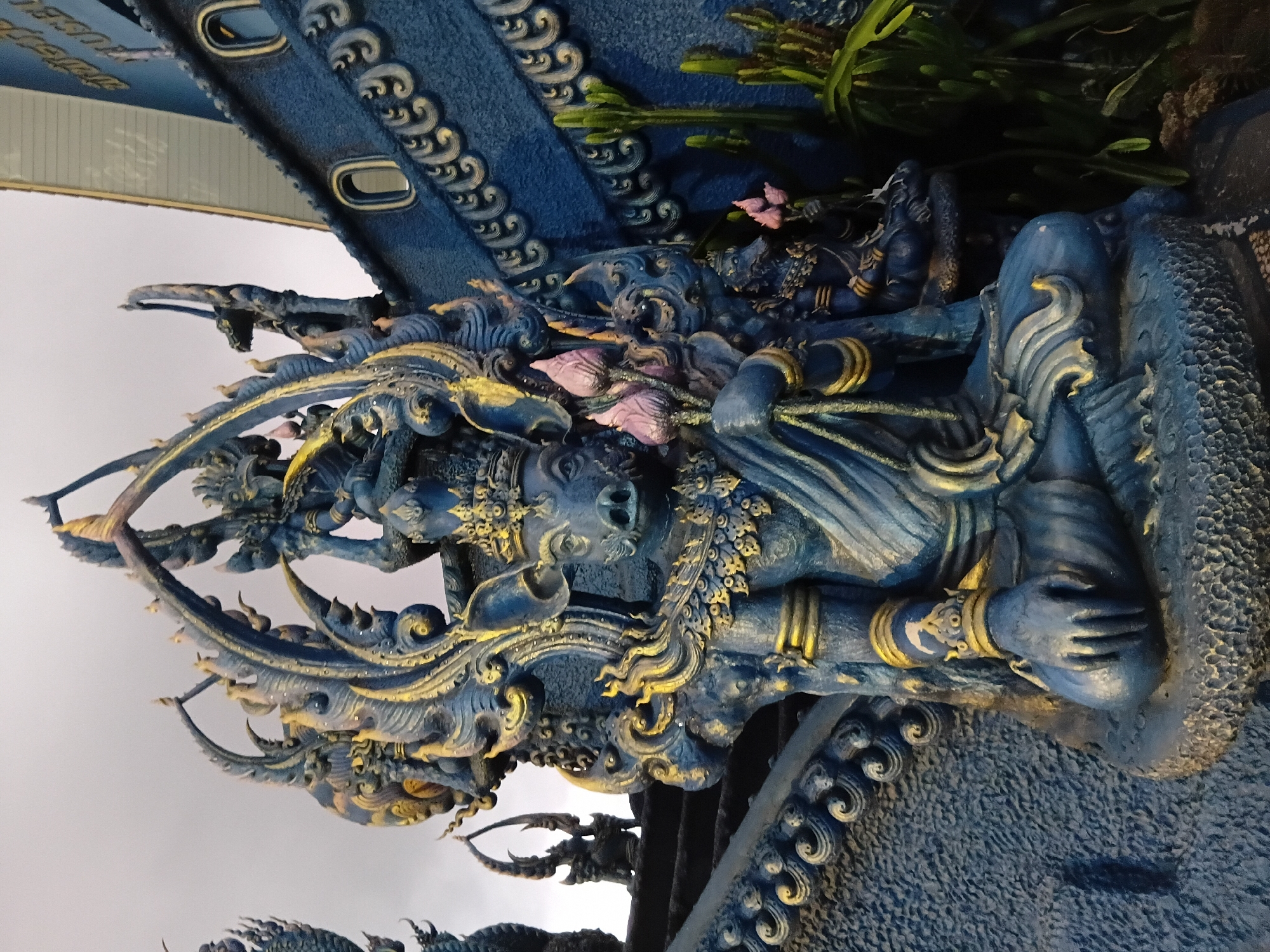 The image captures a detailed view of a statue with a pig's head, adorned with an elaborate headdress and intricate golden embellishments, holding lotus flowers. The statue is crafted from a dark blue material with gold accents. It sits on a textured base, and appears to be part of a larger artistic or architectural installation. Behind the main statue, a blue, boat-like structure with ornate carvings and windows is visible, suggesting it is part of a building. The overall scene is likely located at the Wat Rong Khun (White Temple) in Chiang Rai, Thailand, known for its unique and elaborate Buddhist art. The sky is overcast, and some greenery is present in the foreground.