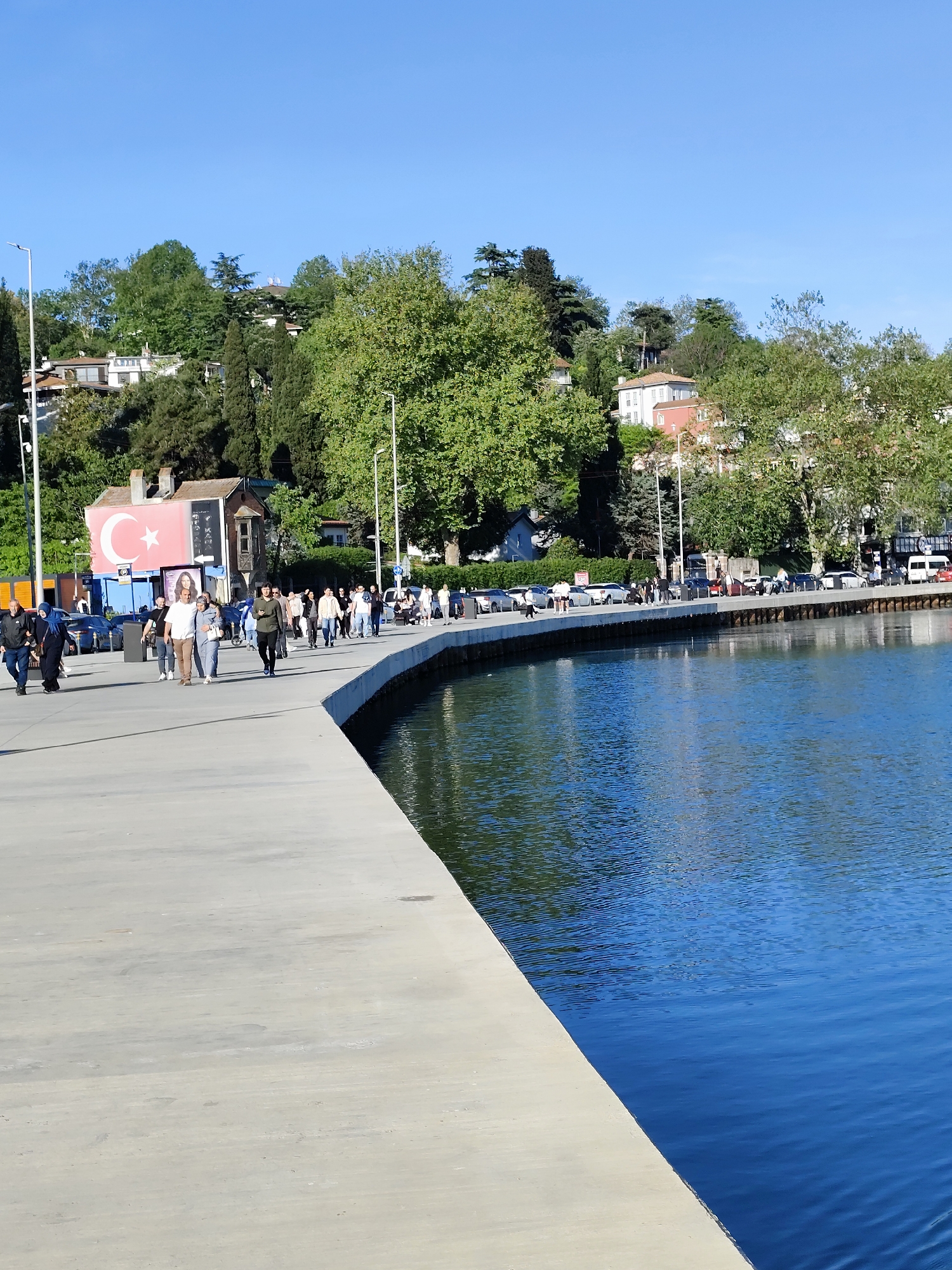 The image depicts a waterfront scene in Sarıyer, Turkey, with a concrete promenade curving along a body of water, likely the Bosphorus. A group of people are walking along the promenade, engaged in casual strolling. Cars are parked along the road that runs adjacent to the walkway. The background features buildings nestled amongst lush greenery on a hillside. A building prominently displays a large Turkish flag with its crescent moon and star. The sky is clear and blue, reflecting in the water.