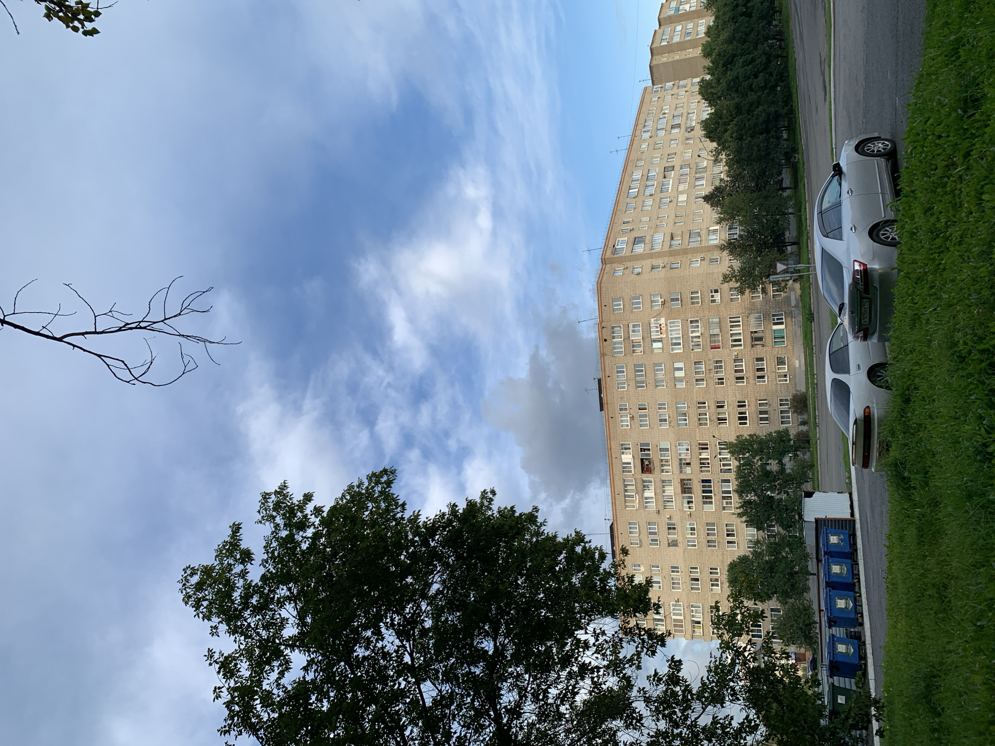 The image depicts a multi-story brick apartment building in Prokopyevsk, Russia. In the foreground, lush green grass covers the ground, with two silver sedans parked on a paved area. Behind the cars, several blue trash receptacles are lined up. A large tree with dark green foliage is positioned to the left, partially obscuring the view of the building. The sky above is a mixture of blue with scattered white and grey clouds. Bare tree branches are visible against the sky in the upper portion of the image. The building itself appears to be a typical Soviet-era apartment block, characterized by its uniform window layout and brick construction.