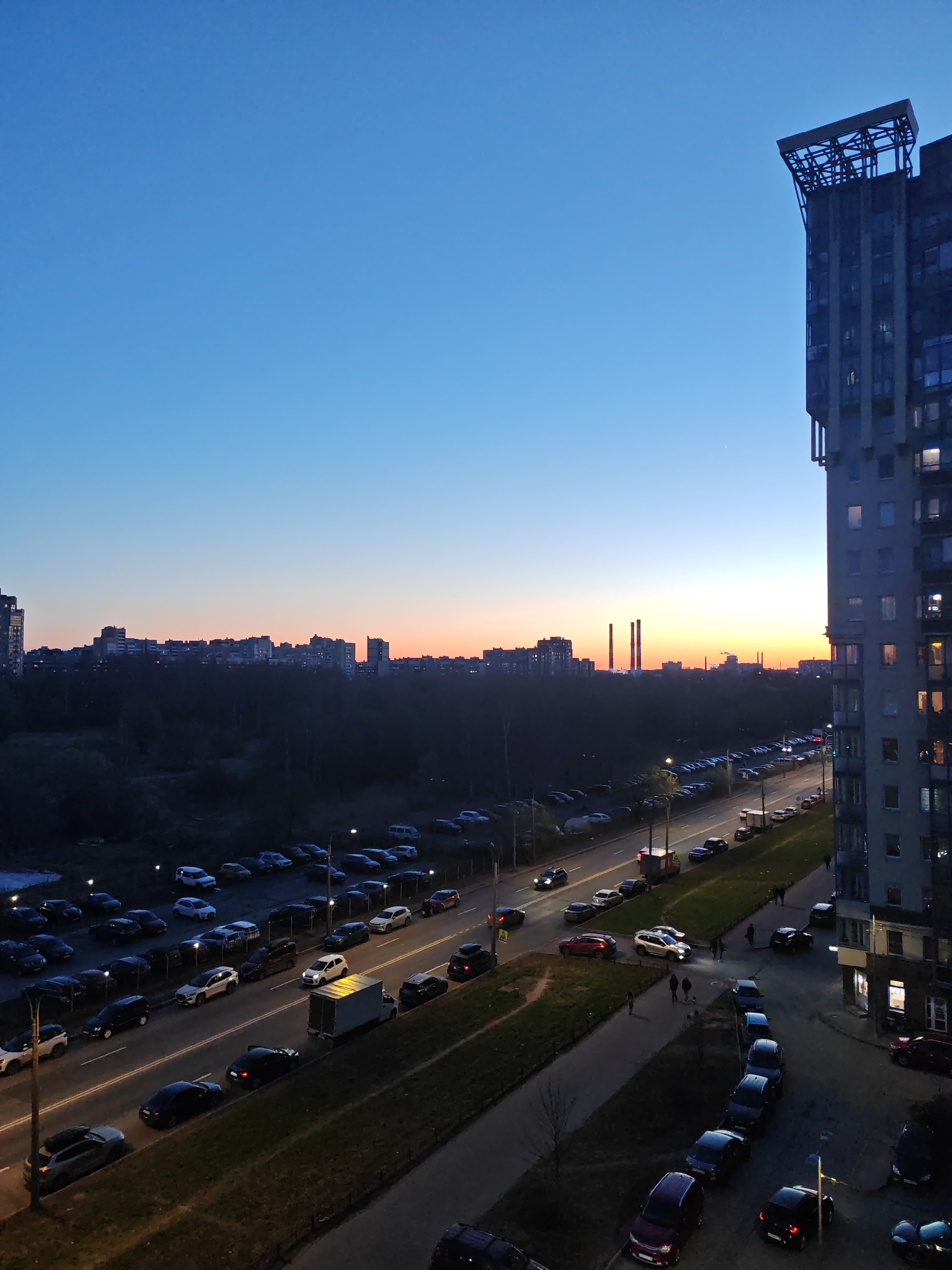 The image shows an urban scene at dusk, likely taken from a high vantage point such as an apartment balcony. The location appears to be Saint Petersburg, Russia, given the architectural style of the buildings and the overall cityscape.
The sky transitions from a deep blue at the top to a warm orange-yellow near the horizon, indicating either sunrise or sunset. In the background, a silhouette of buildings and trees forms the skyline. Two prominent industrial chimneys are visible, possibly belonging to a power plant.
Below the skyline is a dense area of trees, obscuring much of the ground. In the middle ground, a wide street runs across the frame. On the left side of the street, there is a parking lot filled with many cars. Cars are also parked along the right side of the street. Traffic is flowing in both directions.
Pedestrians are seen walking on the sidewalk adjacent to the road on the right side of the frame. One group of people is crossing the street.
On the right of the frame stands a tall, multi-story apartment building, obscuring parts of the scene, and with a prominent architectural structure at the top. The building is lit by interior lights in some windows, and the overall lighting is dim due to the time of day.