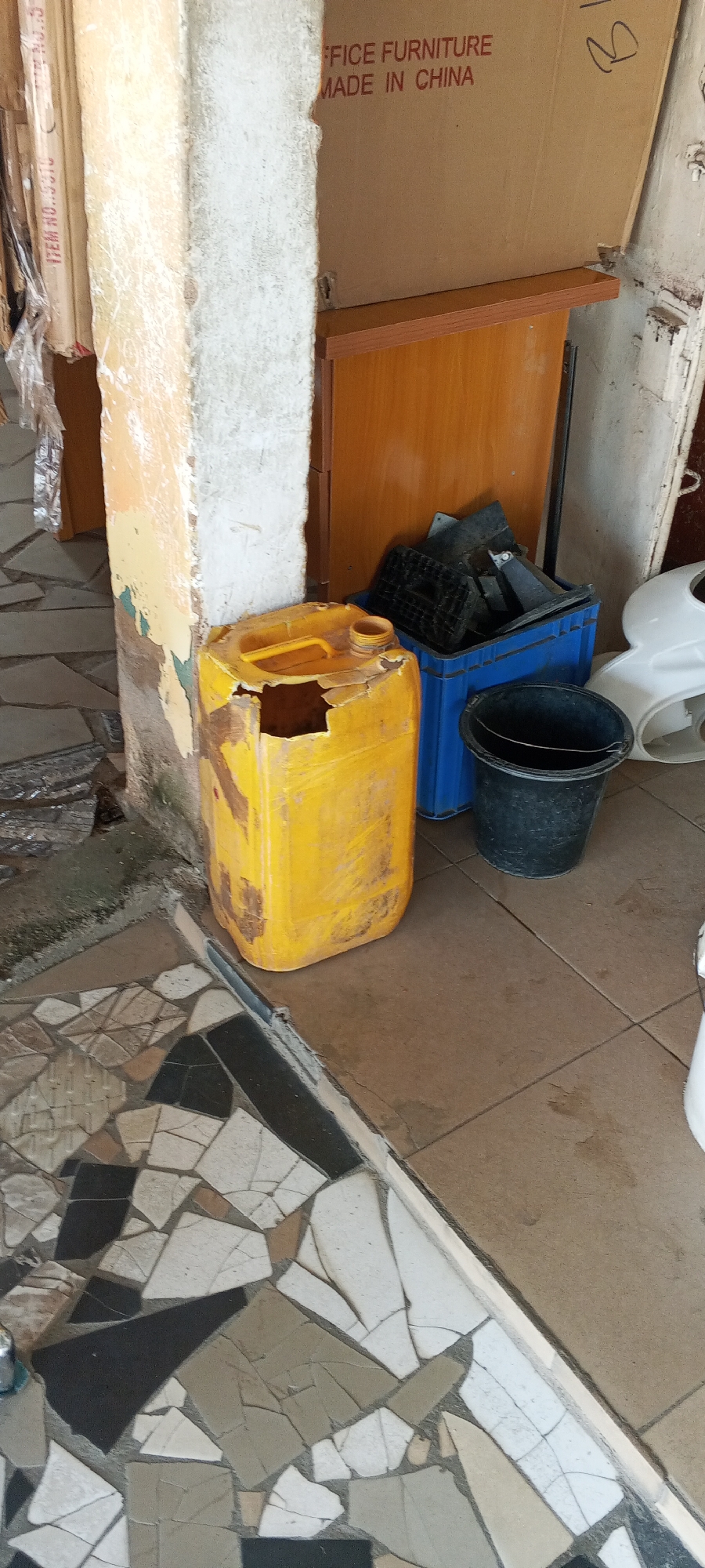 The image captures an outdoor scene in Jalingo, Nigeria, likely a storage area or backyard. In the foreground, a mosaic tiled floor with a white and brown pattern transitions to a plain brown tiled floor. To the left, a weathered, peeling yellow plastic jerrycan with a large, broken opening sits on the brown tiles. Behind the jerrycan, a rough, light-colored concrete pillar stands, showing signs of age and damage, with portions of yellow and green paint peeling off.
Further into the scene, to the right of the pillar, a large, open cardboard box with red text that reads "FFICE FURNITURE MADE IN CHINA" is propped up. Beneath the box, a light brown wooden cabinet or shelf is visible. Next to the jerrycan, a bright blue plastic crate is filled with various black plastic items, possibly broken parts or containers. In front of the blue crate sits a black plastic bucket with a metal handle. Partially visible on the far right is a white object, possibly part of a toilet or sink, suggesting a potential outdoor plumbing supply area. The overall environment appears utilitarian and somewhat cluttered, with elements indicative of a workshop or storage space.