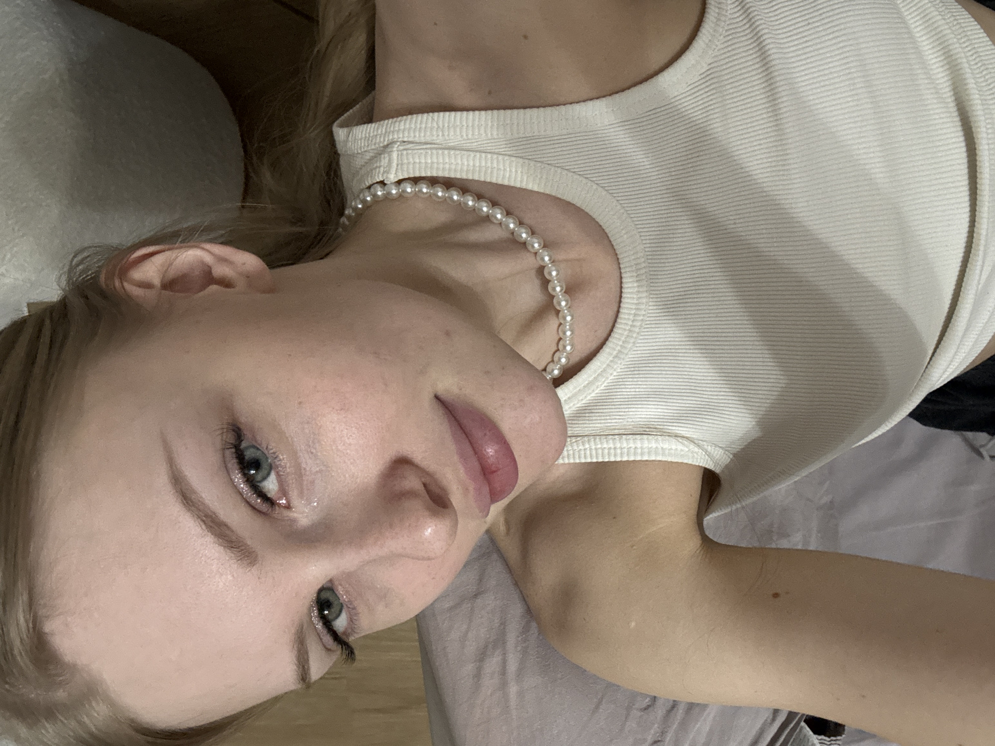 A young woman is depicted in an overhead, rotated shot, lying on a soft, light-colored surface, possibly a bed. Her head is positioned towards the left side of the frame, and she looks directly into the camera with a neutral to slightly pensive expression. She has fair skin, blonde or light brown hair that flows around her, and piercing blue-green eyes. Her eyes are enhanced with what appear to be long, dark eyelashes and a subtle shimmer or glitter on her lower lash line, while her eyebrows are neatly defined. Her lips are full and slightly parted, appearing to have a natural or light pink gloss.
She is wearing a simple white ribbed tank top, and a single strand of pearl beads forms a necklace around her neck. The ribbed texture of the tank top is prominent. Portions of her bare arm and shoulder are visible, confirming her fair complexion and revealing a small mole on her upper arm. The background is indistinct but appears to be indoor, consistent with a bedroom setting, featuring muted tones of fabric or bedding. The image focuses closely on the woman, with no other prominent objects or activities discernible. No text is visible in the media. This scene is located in Perm, Russia.