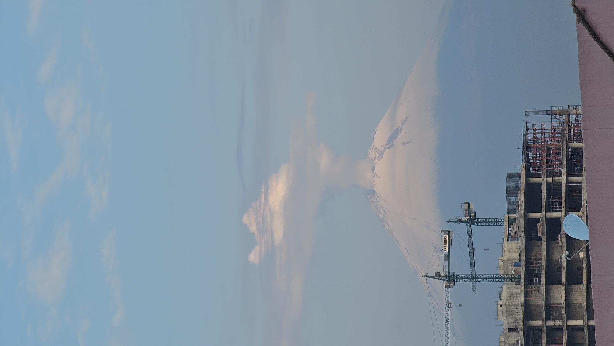 The image captures a panoramic view of an urban landscape with a prominent natural landmark in the background, likely during early morning or late afternoon. The scene is dominated by a clear, light blue sky scattered with soft white and light pink clouds, suggesting either dawn or dusk.
In the foreground, on the right side, a partial view of an existing building with a reddish-brown exterior wall is visible. A blue satellite dish is mounted on this wall, and a rope or cable runs along its top edge.
Adjacent to this, in the mid-ground, a large construction site is evident. An unfinished multi-story building, constructed with concrete and rebar, shows signs of ongoing work with scaffolding structures. Two prominent green tower cranes are active at the site; the boom of one crane clearly displays the text "ESPAMEX."
Dominating the background on the left is a majestic, snow-capped or ice-covered mountain, characteristic of a volcano, with a distinct conical peak. From near its summit, a light-colored plume, likely composed of ash or steam, rises into the sky and extends horizontally as a cloud formation. Given the location context of Puebla, Mexico, this mountain is identifiable as Popocatépetl, an active volcano known for its frequent emissions.
The image effectively contrasts human development and construction with the powerful and active forces of nature.