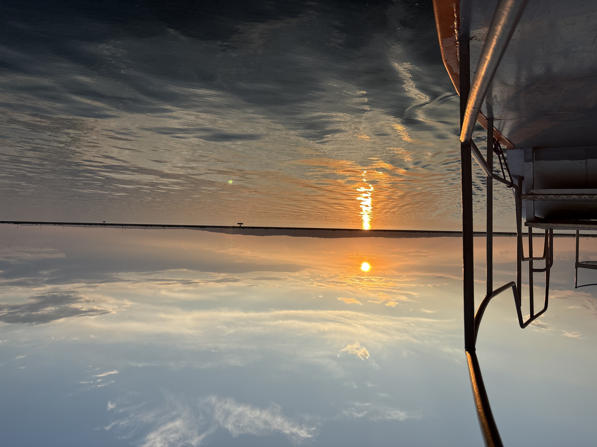 The image depicts a serene sunset over a body of water, likely the Kama River near Chaikovsky, Russia. In the foreground, the railing and deck of a boat are visible, suggesting the vantage point is from aboard a vessel. The sun, a bright orb, is low on the horizon, casting a golden reflection across the rippling water. The sky is a gradient of soft blues, oranges, and yellows, with scattered wispy clouds. In the distance, a faint silhouette of the land can be observed, possibly the shoreline of Chaikovsky, with structures or trees lining the horizon. A few small boats or buoys are visible on the water. The overall atmosphere is peaceful and tranquil, characteristic of a sunset view.