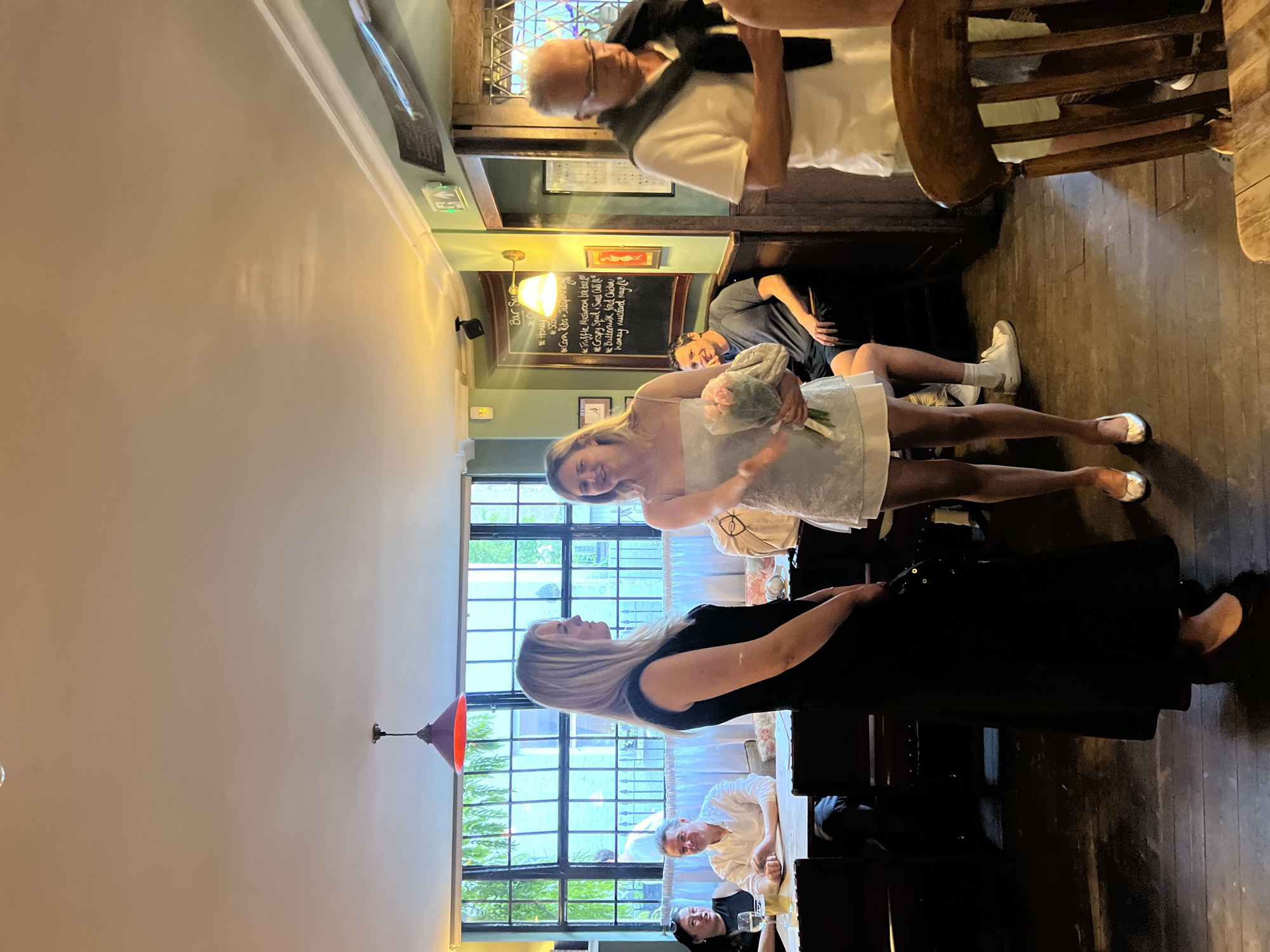 The image depicts a group of people in what appears to be a restaurant or pub. The location seems to be in London, United Kingdom, based on the architectural style of the windows and the general ambiance.
A woman in a black dress is standing, engaging in conversation with another woman who is holding a bouquet of flowers and wearing a light-colored dress, possibly a wedding dress. There are several other individuals visible in the background: a man in a white shirt and grey cardigan, a younger man sitting in a booth, and two women seated at a table by the window.
The interior includes wooden floors, a chalkboard menu, and traditional-style lighting fixtures. The overall atmosphere is lively and social.