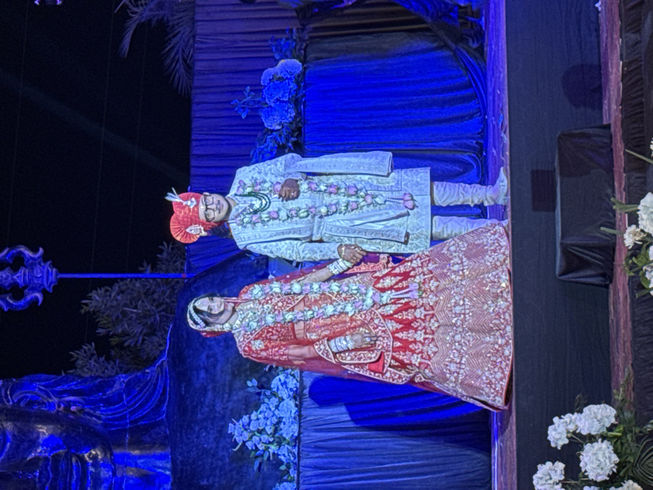 A wedding scene presents a bride and groom in traditional attire, adorned with floral garlands. The groom wears a patterned sherwani and a turban, while the bride is in a red lehenga. They stand before a backdrop of blue fabric and floral arrangements, under lighting that casts a blue hue. The scene is elaborately decorated, with elements suggesting a staged setting.