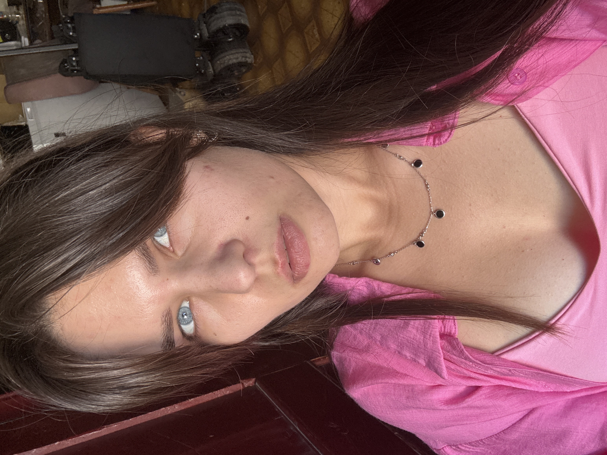 A medium shot shows a young woman with long, dark brown hair and blue eyes. She is wearing a pink top and a silver necklace with black pendants. Her head is slightly tilted to the left, and she is looking directly at the camera. The background is out of focus, but it appears to be an indoor setting with some objects that look like furniture and appliances. A dark red wooden surface is visible on the left side of the image. The lighting is soft and natural.