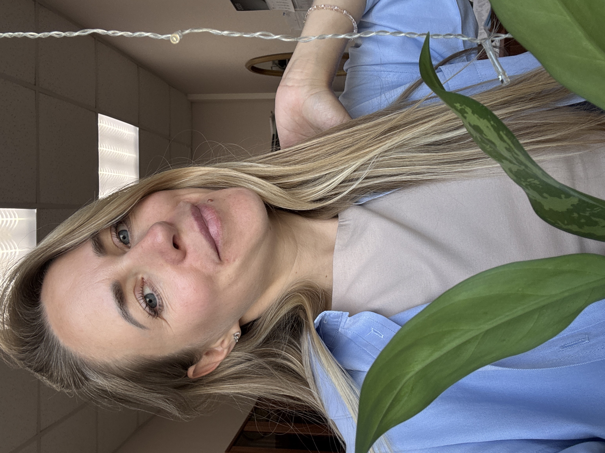 A woman with long blonde hair and blue eyes is photographed from the chest up. She is wearing a light blue collared shirt over a beige top. Her right hand is raised to her neck. She is looking towards the camera with a slight smile. A plant with large green leaves is partially visible in the foreground, obscuring the lower portion of the image. In the background, a ceiling with fluorescent lighting panels is visible, as well as a wall with a calendar and a mirror. The scene appears to be indoors, possibly an office or home environment. There is no indication of the location being in Chaikovsky, Russia.