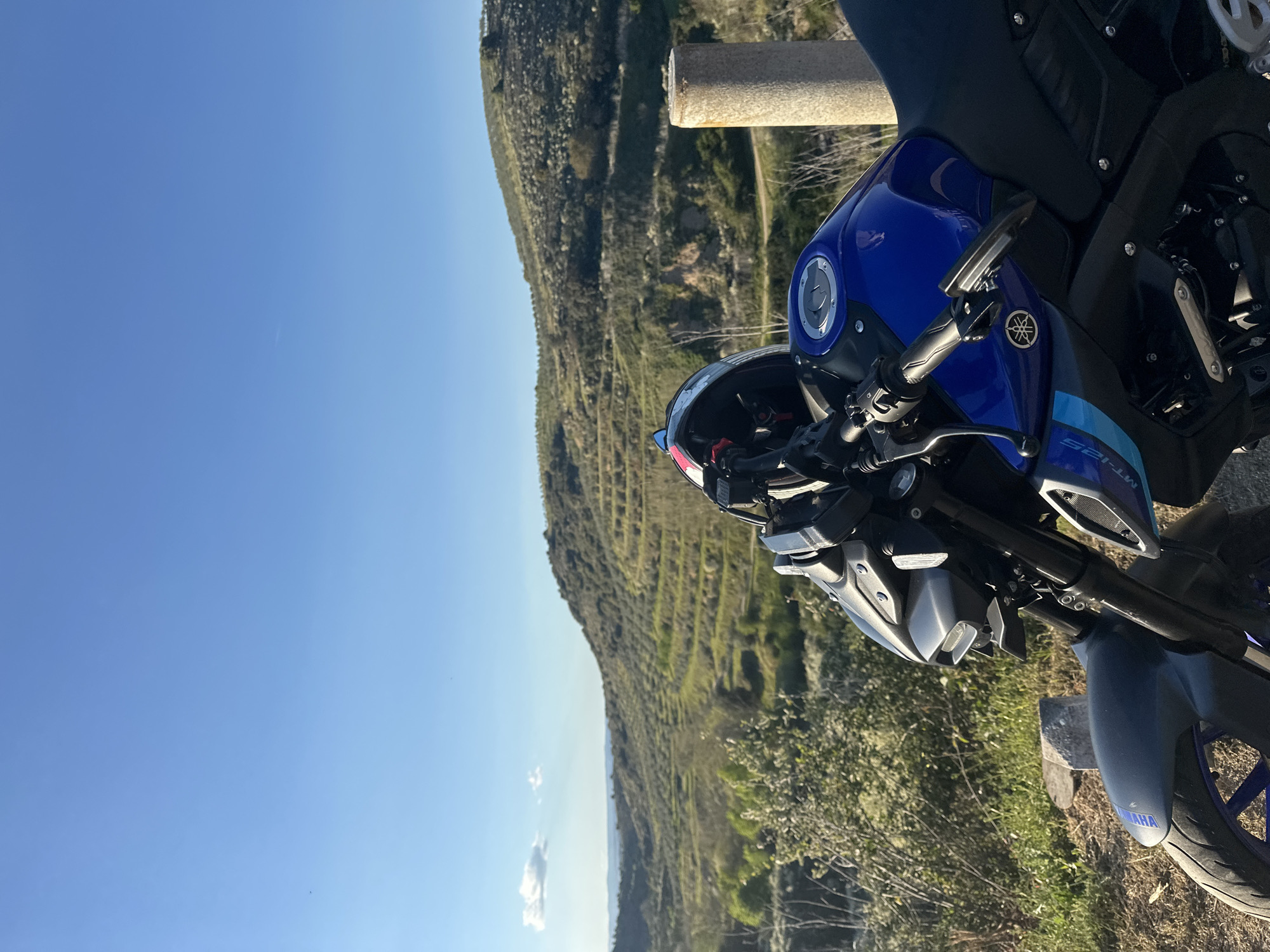 A blue Yamaha MT-125 motorcycle is parked on a roadside in a rural area near Mirandela, Portugal. The motorcycle's front wheel and blue fuel tank are visible, along with the handlebars and a motorcycle helmet resting on them. The background features a mountainous terrain covered with green vegetation and a clear blue sky. A concrete roadside barrier is positioned near the motorcycle. The photograph is taken at an angle.